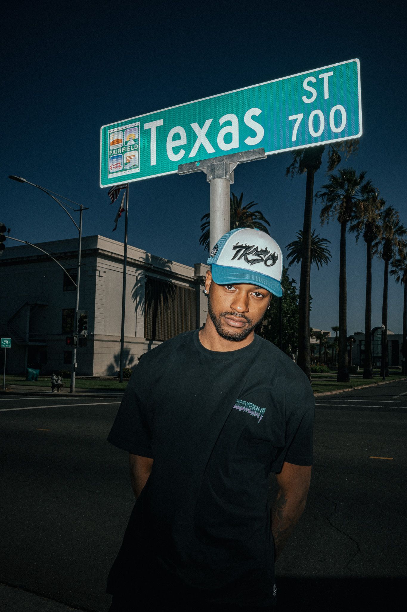 Man in black tee and blue cap stands under Texas St 700 sign with palm trees at dusk.