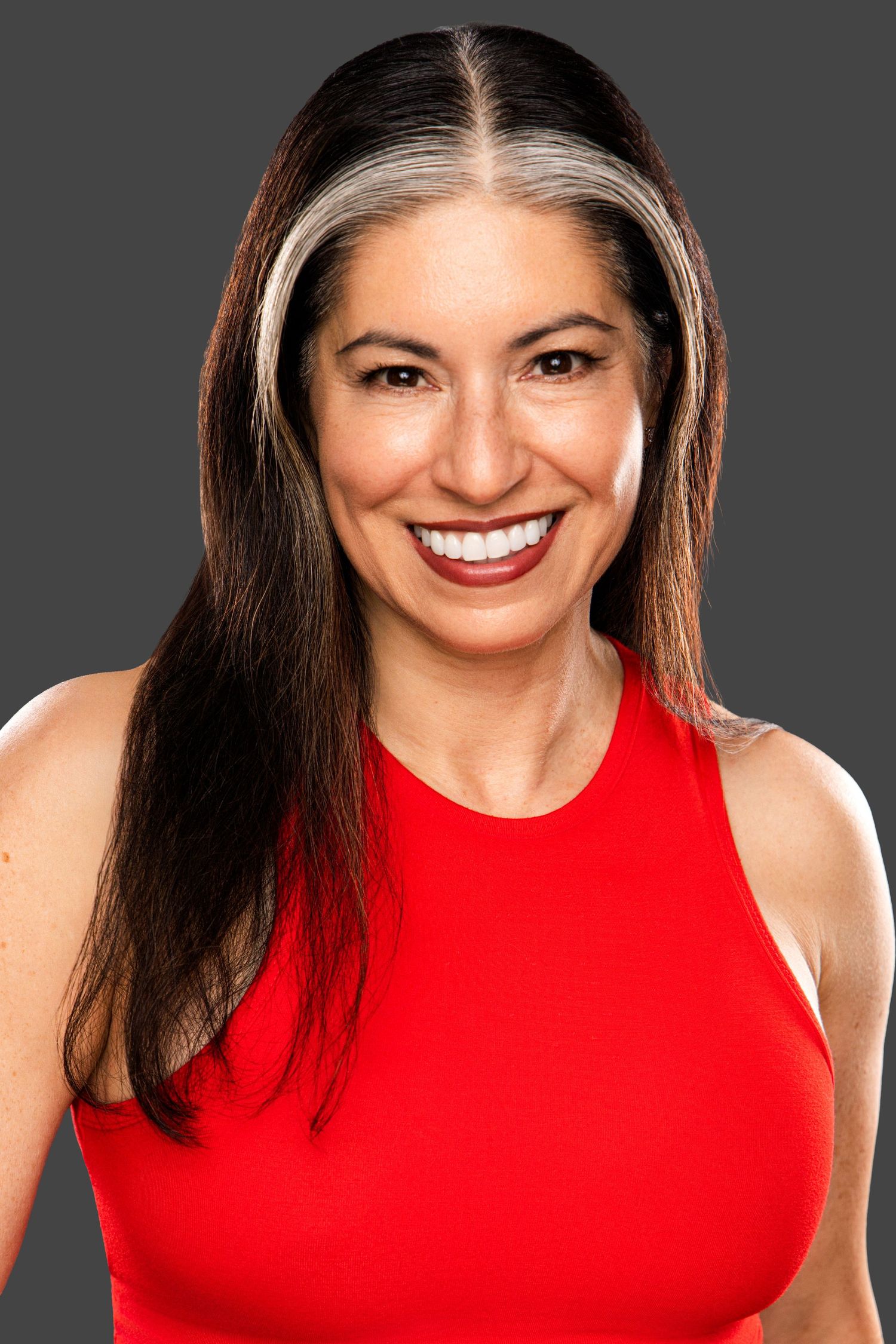 Smiling woman with dark hair and silver highlights wearing a red sleeveless top against a gray background.