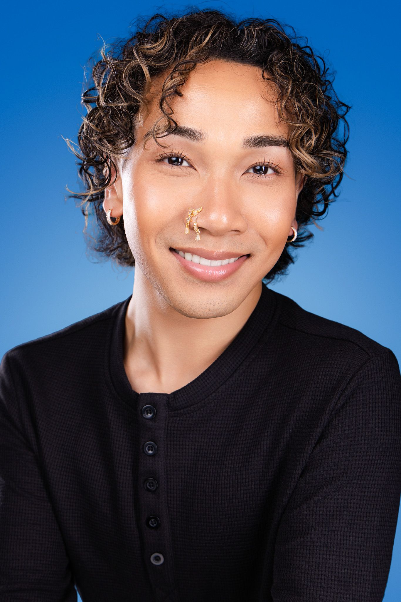 Smiling person with curly hair and gold nose ring wearing black henley shirt against blue background.