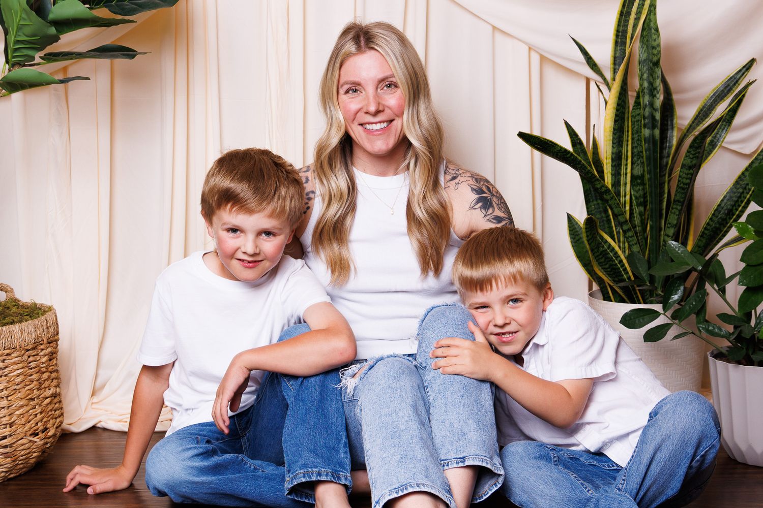 Smiling blonde woman sitting on floor with two young boys in white shirts and jeans, surrounded by green plants.