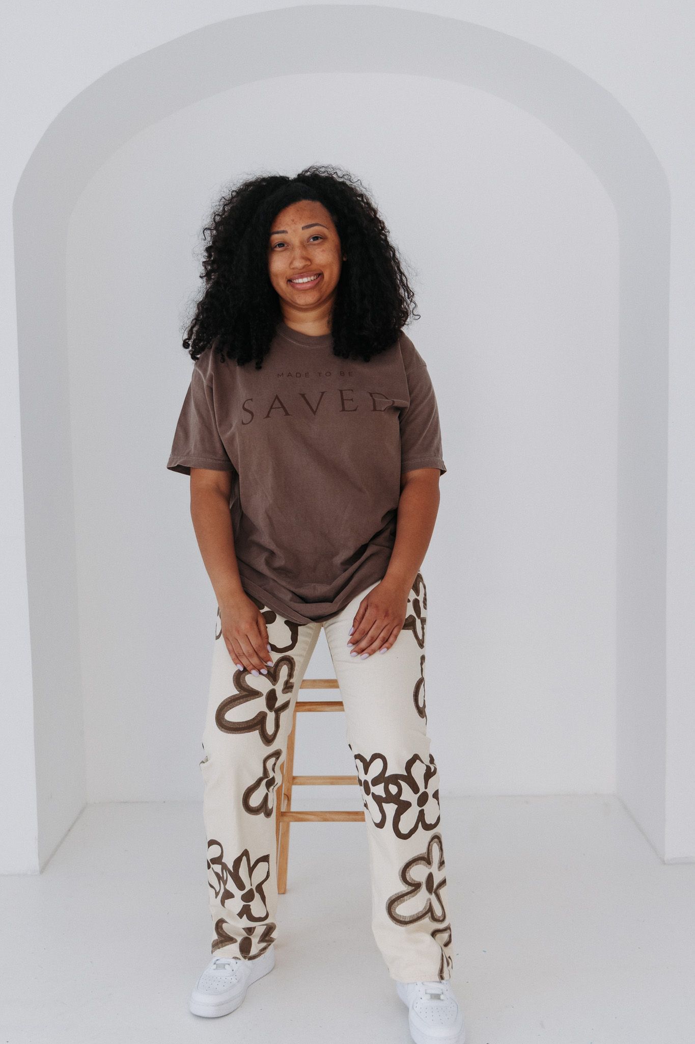 Model seated on wooden chair wearing brown t-shirt and patterned pants in minimalist white setting.