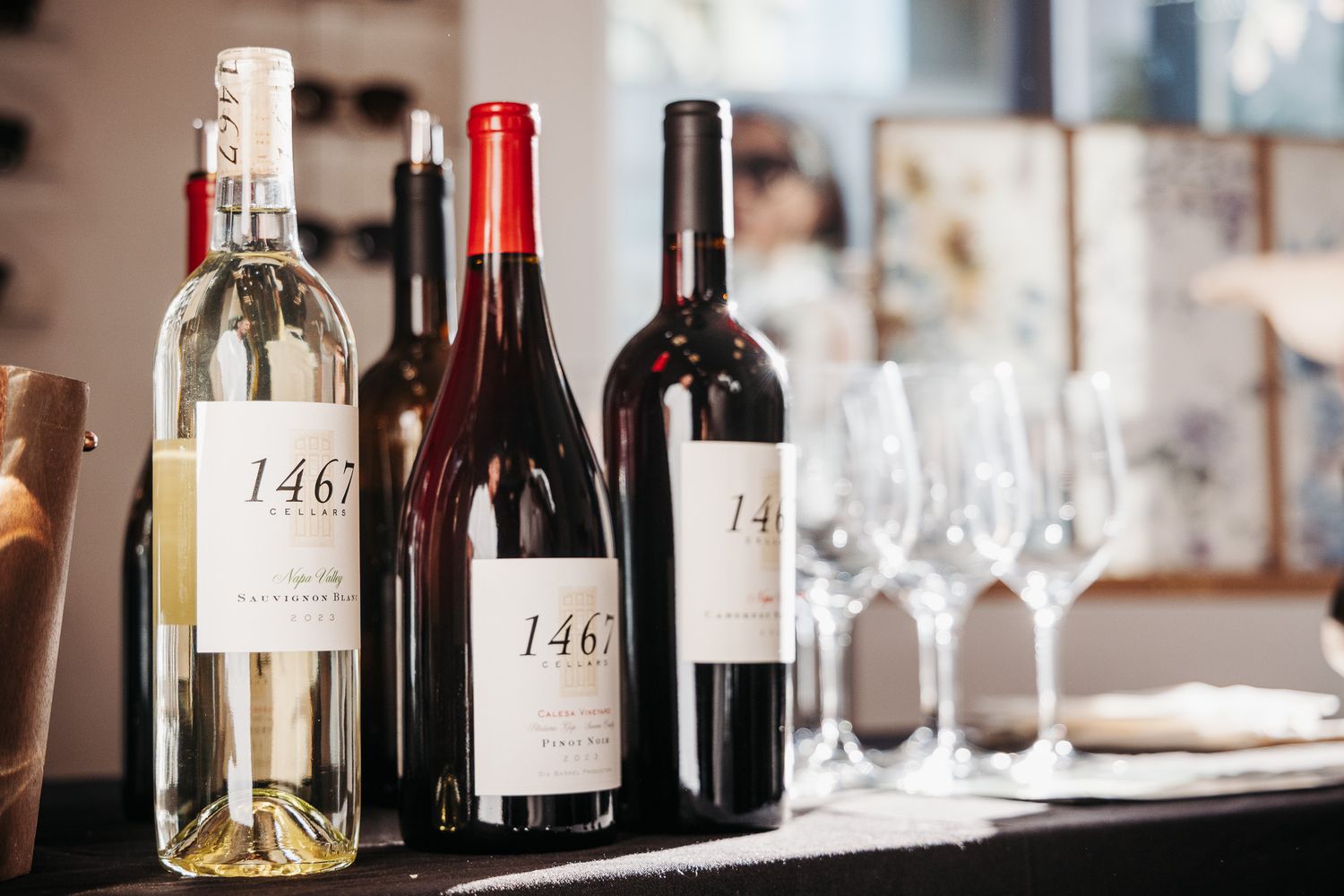 A collection of wine bottles and glasses lined up on a rustic bar counter in soft lighting.