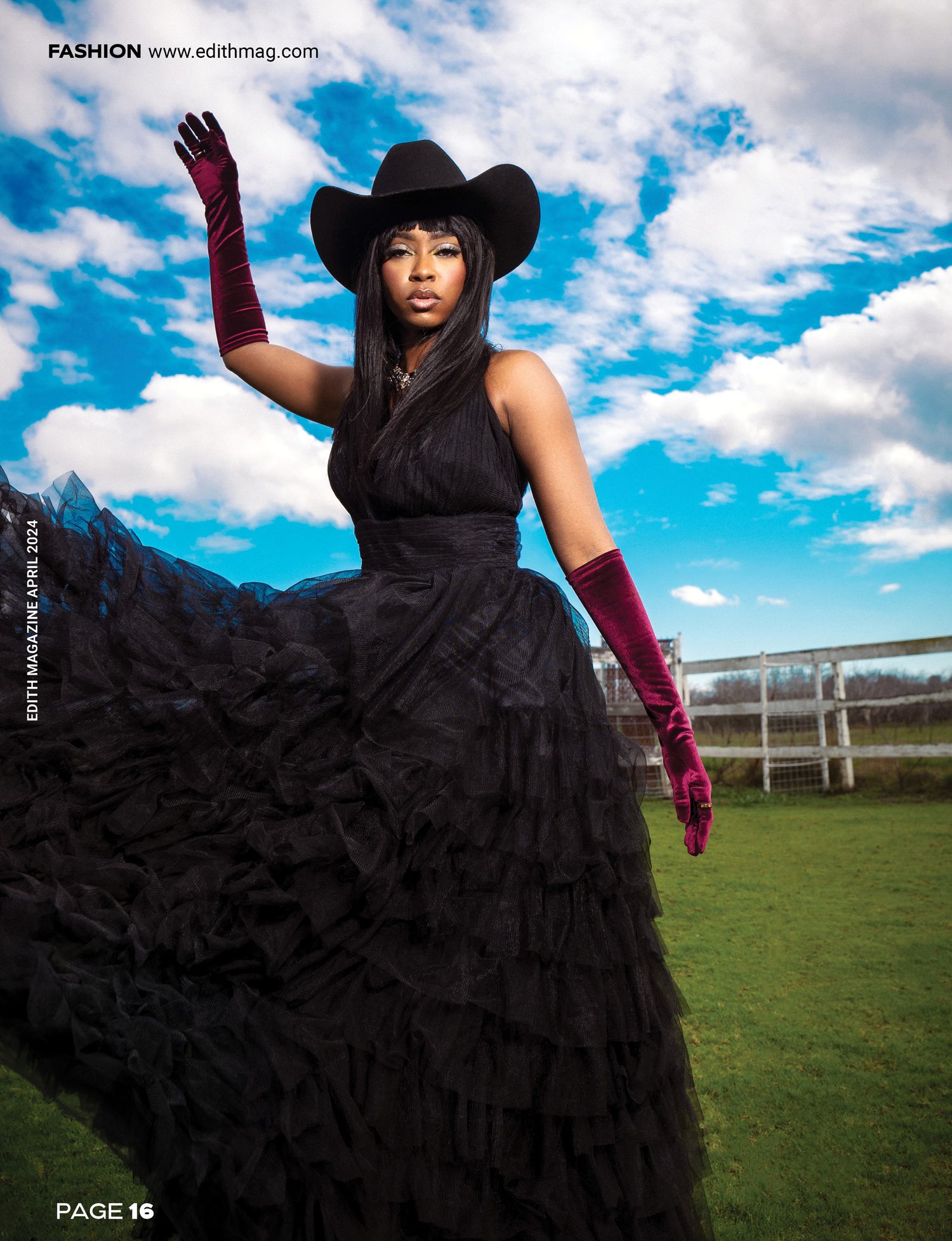 Model in black ruffled gown and cowboy hat poses dramatically with red gloves against cloudy mountain landscape.
