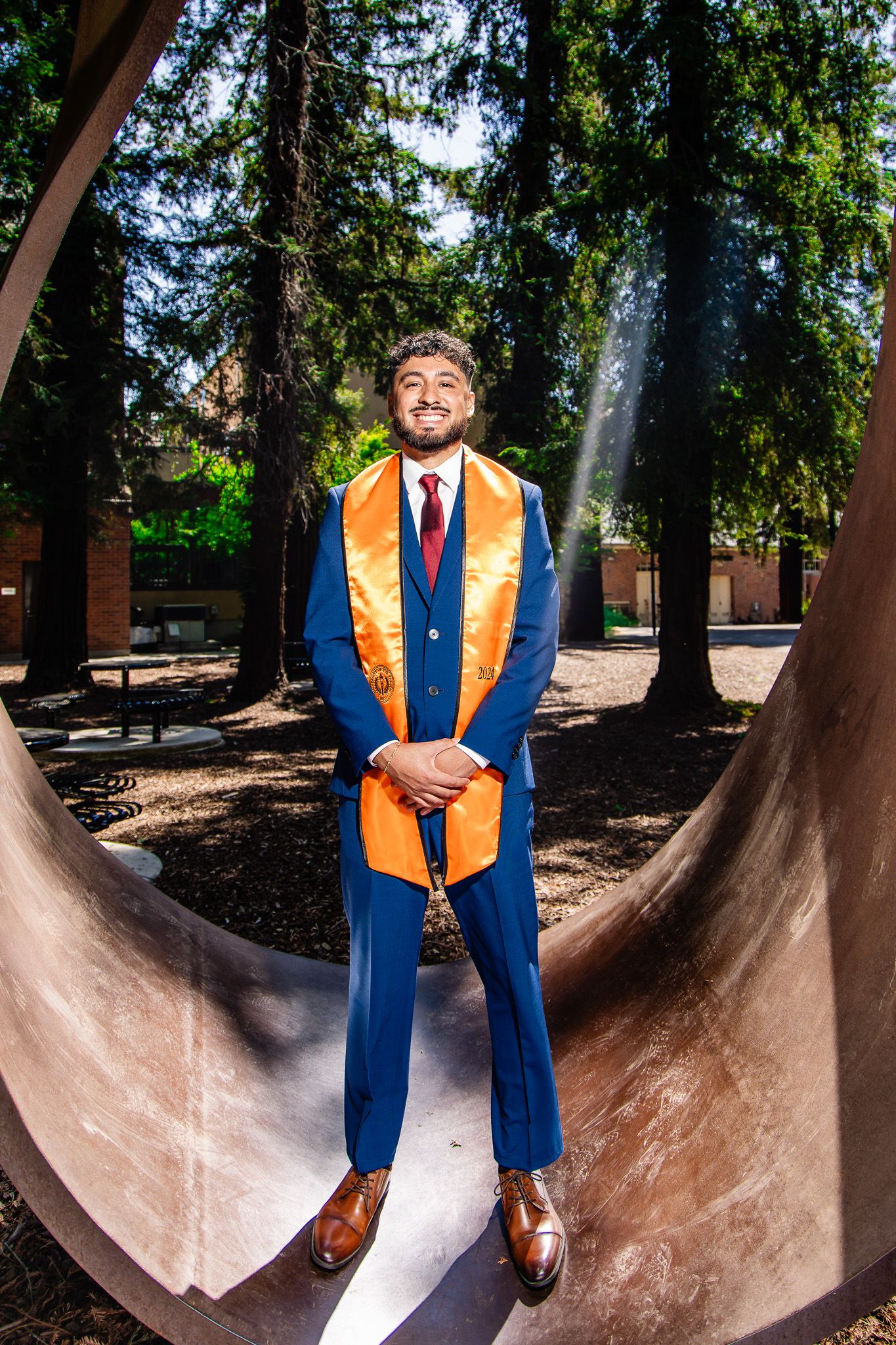 Graduate in blue suit and orange graduation stole poses on curved sculpture outdoors among tall trees.