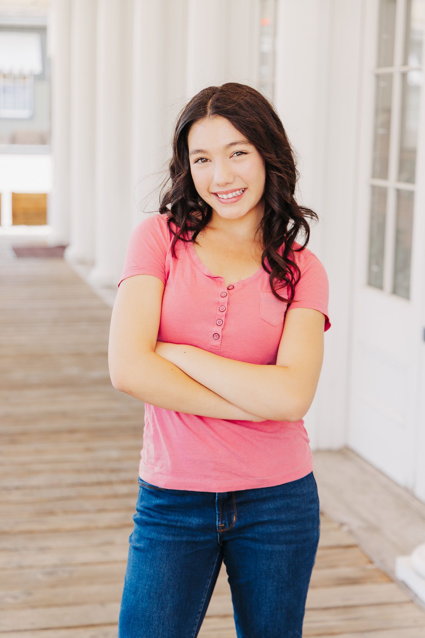 Person wearing a pink button-up short sleeve top and blue jeans poses with arms crossed on a covered porch.