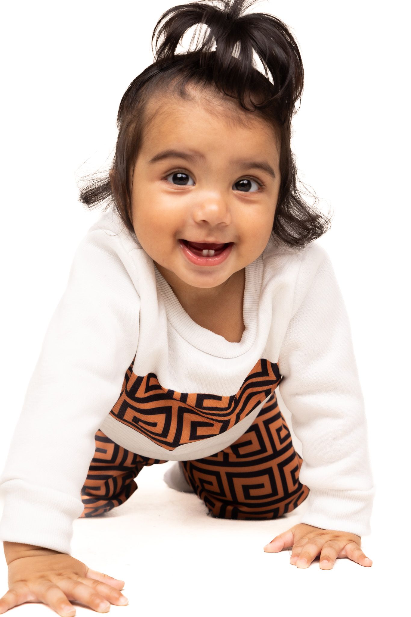 Baby in white and brown patterned outfit smiling and crawling on white background.