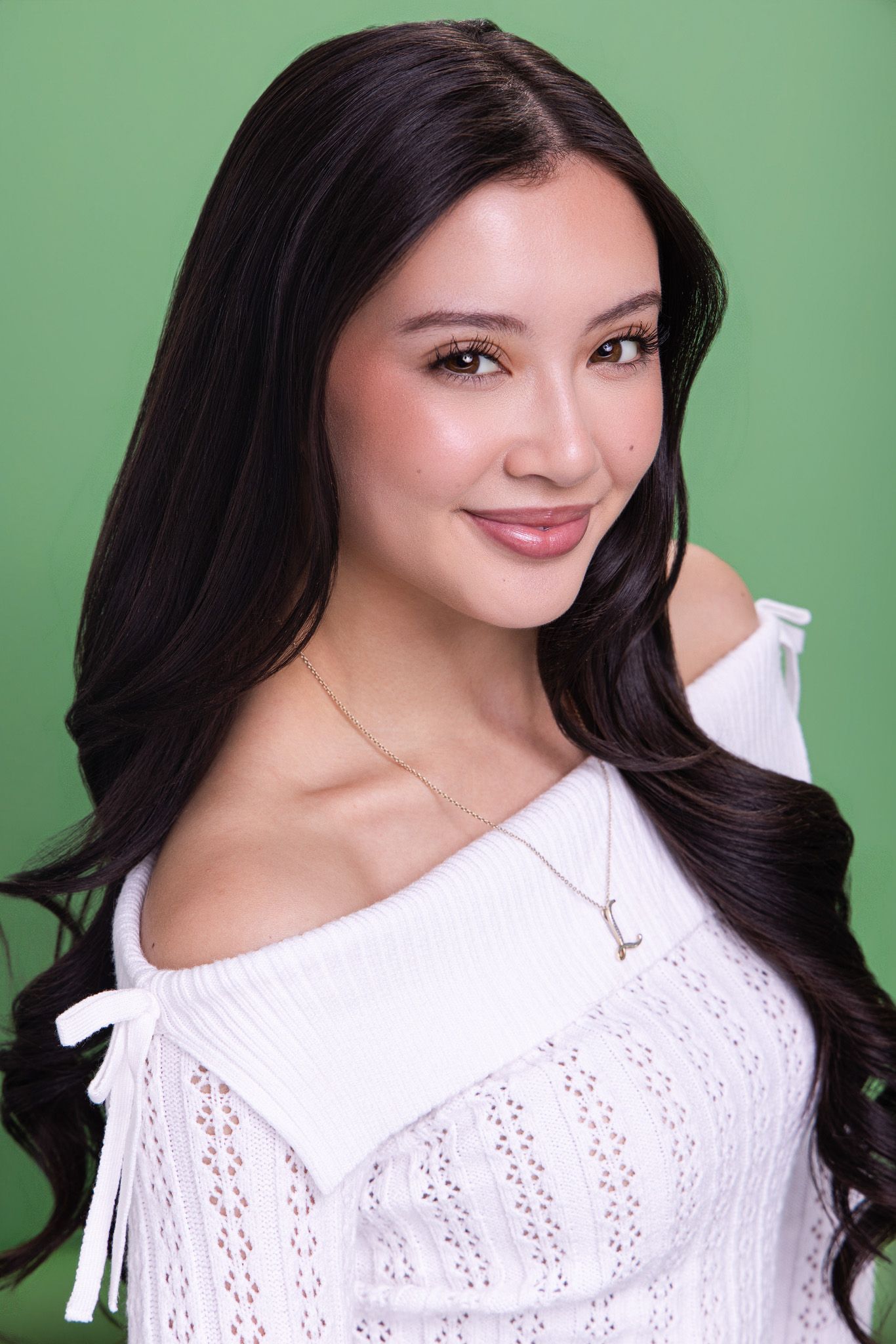 Elegant woman with long dark hair in white off-shoulder top and gold necklace against green background.