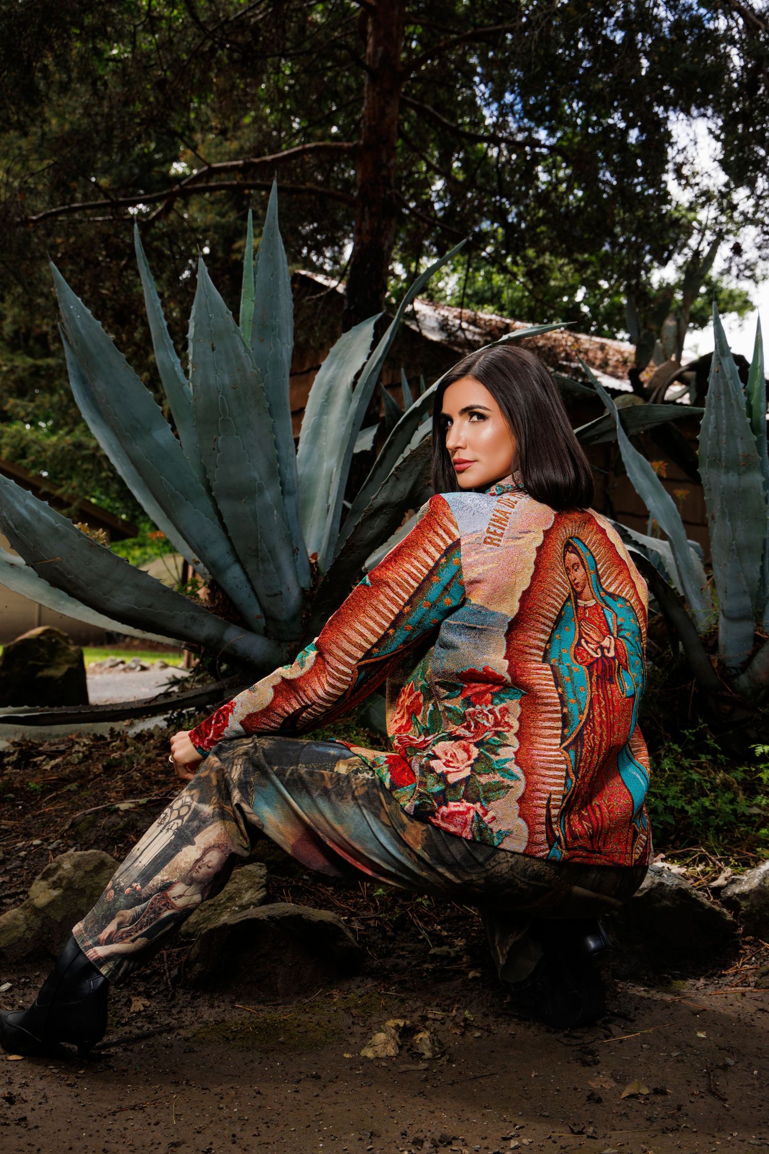 Woman in colorful floral printed coat sits among agave plants in a lush outdoor garden setting.
