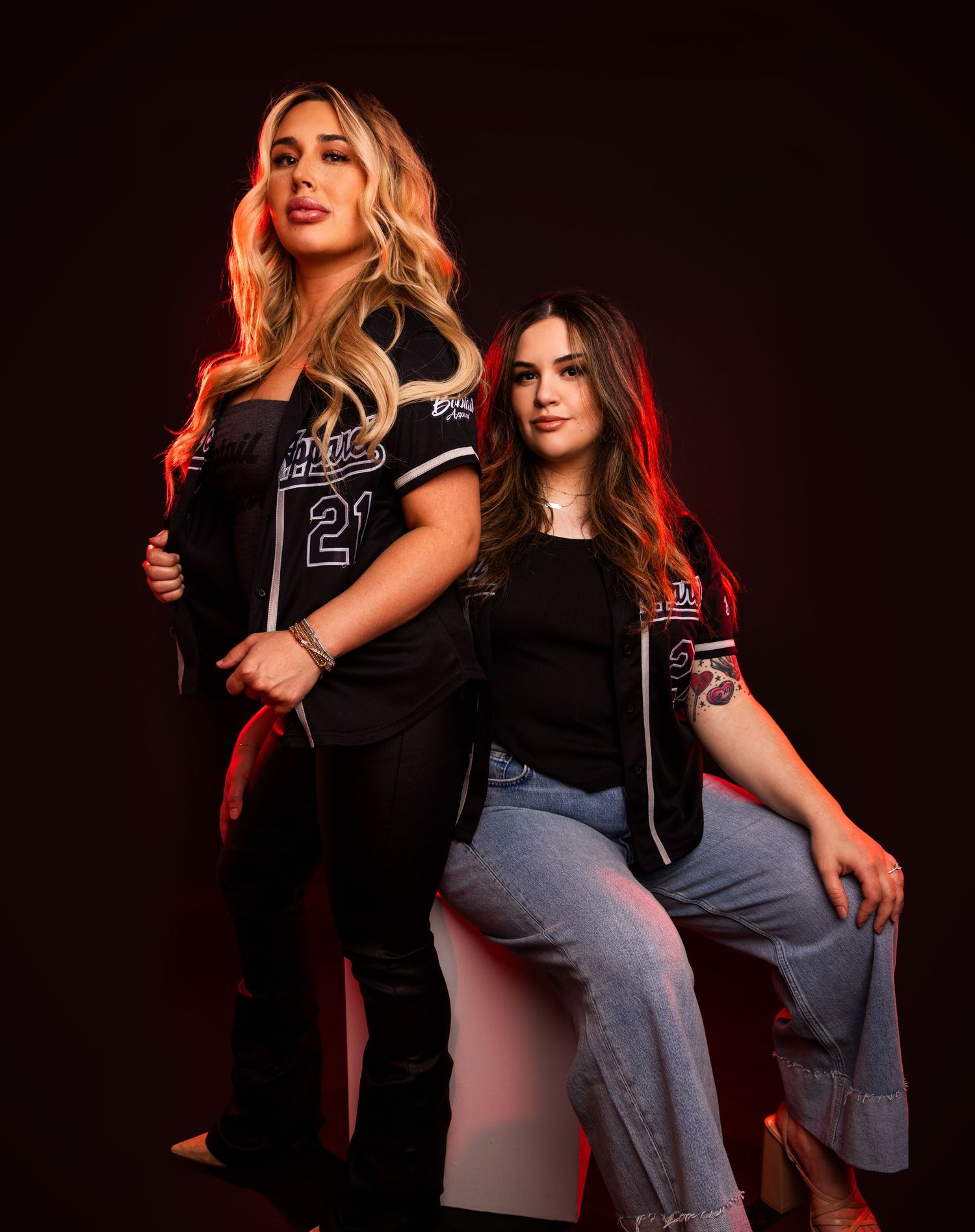 Two models pose together in black and gray outfits against a dark background in a fashion photoshoot series.