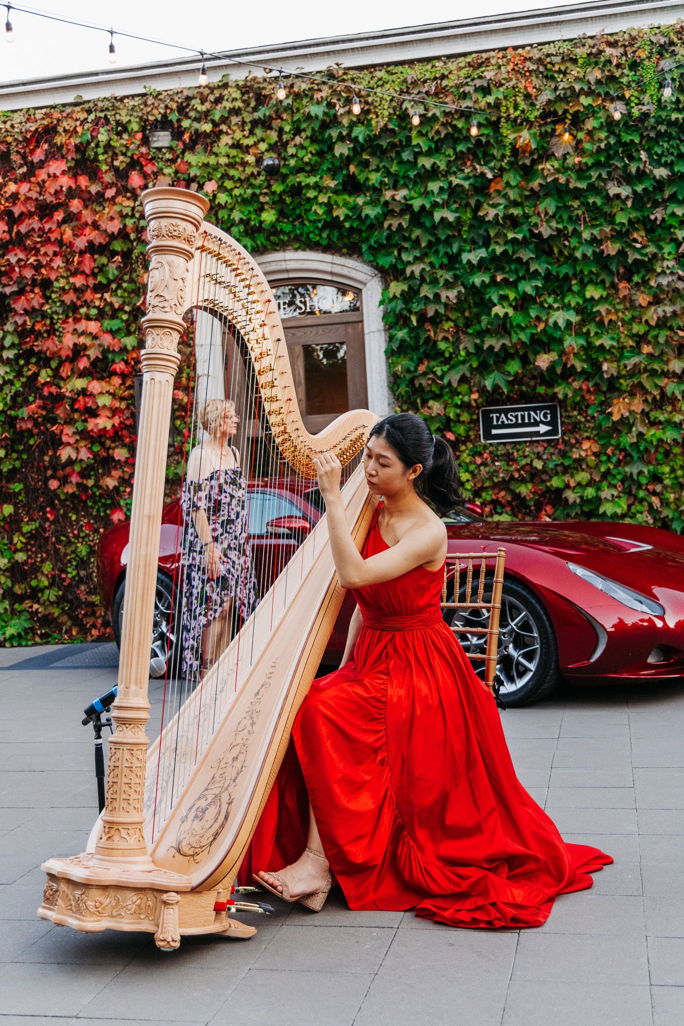 A musician in a flowing red gown performs on an elegant golden harp in an outdoor courtyard setting.