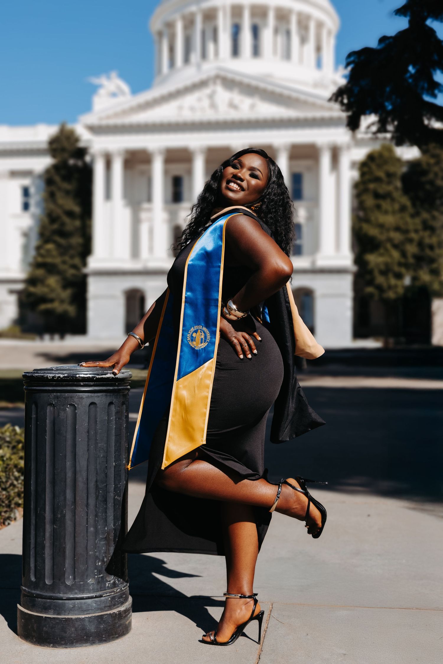 A graduate poses for celebratory photos in front of a government building with white columns and domed architecture.