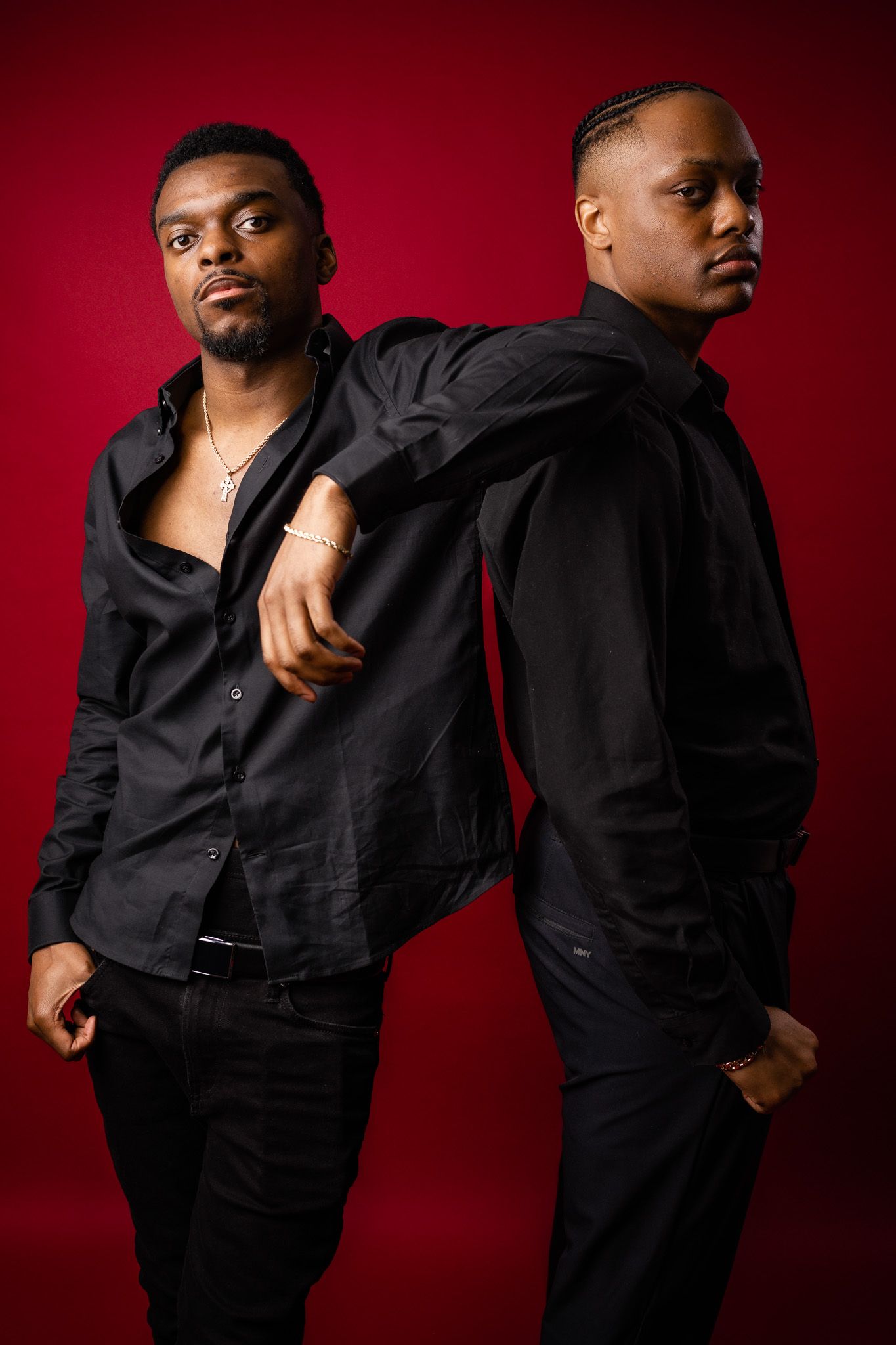 Two men pose back-to-back wearing black outfits against a red background in a dramatic studio portrait.