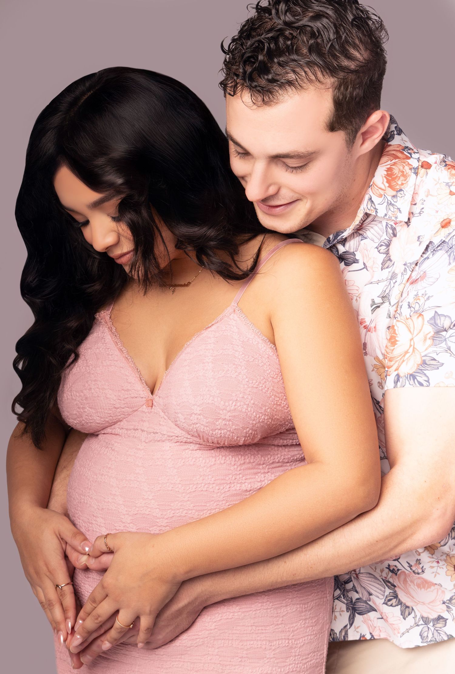 Expectant couple shares intimate moment in studio photo with woman wearing pink dress and man in floral shirt.
