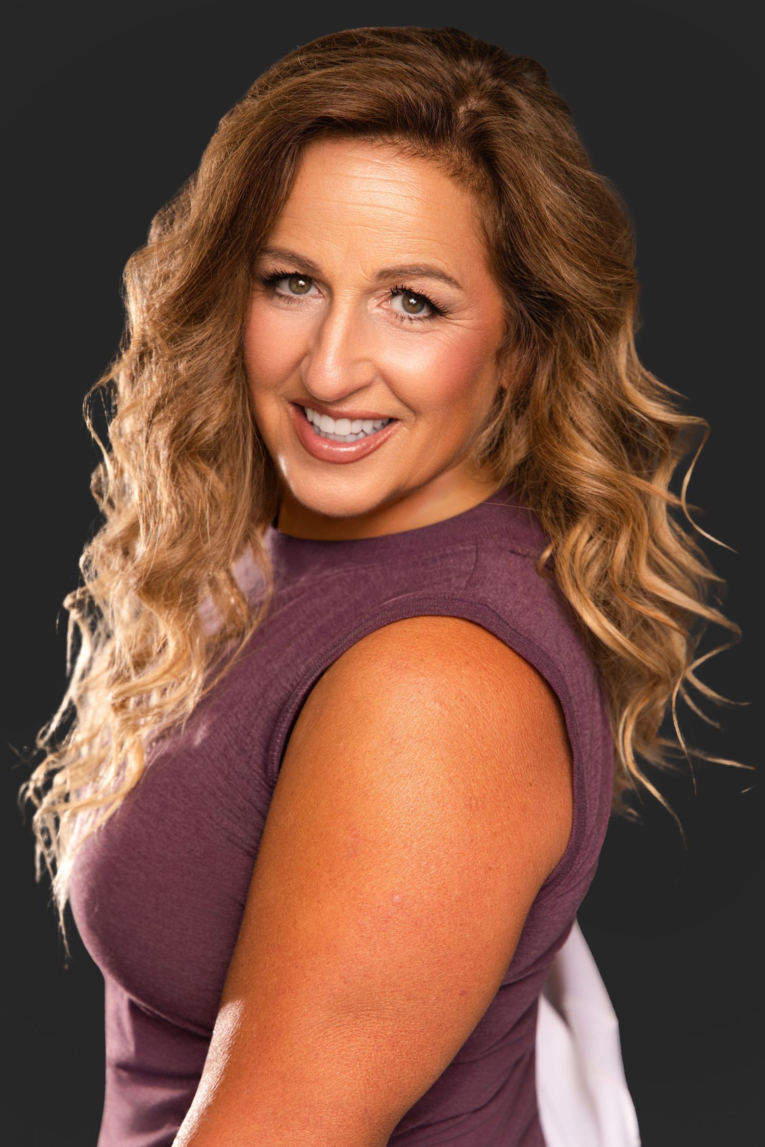 Smiling woman with curly blonde hair wearing a purple sleeveless top, posing against a dark gray background.