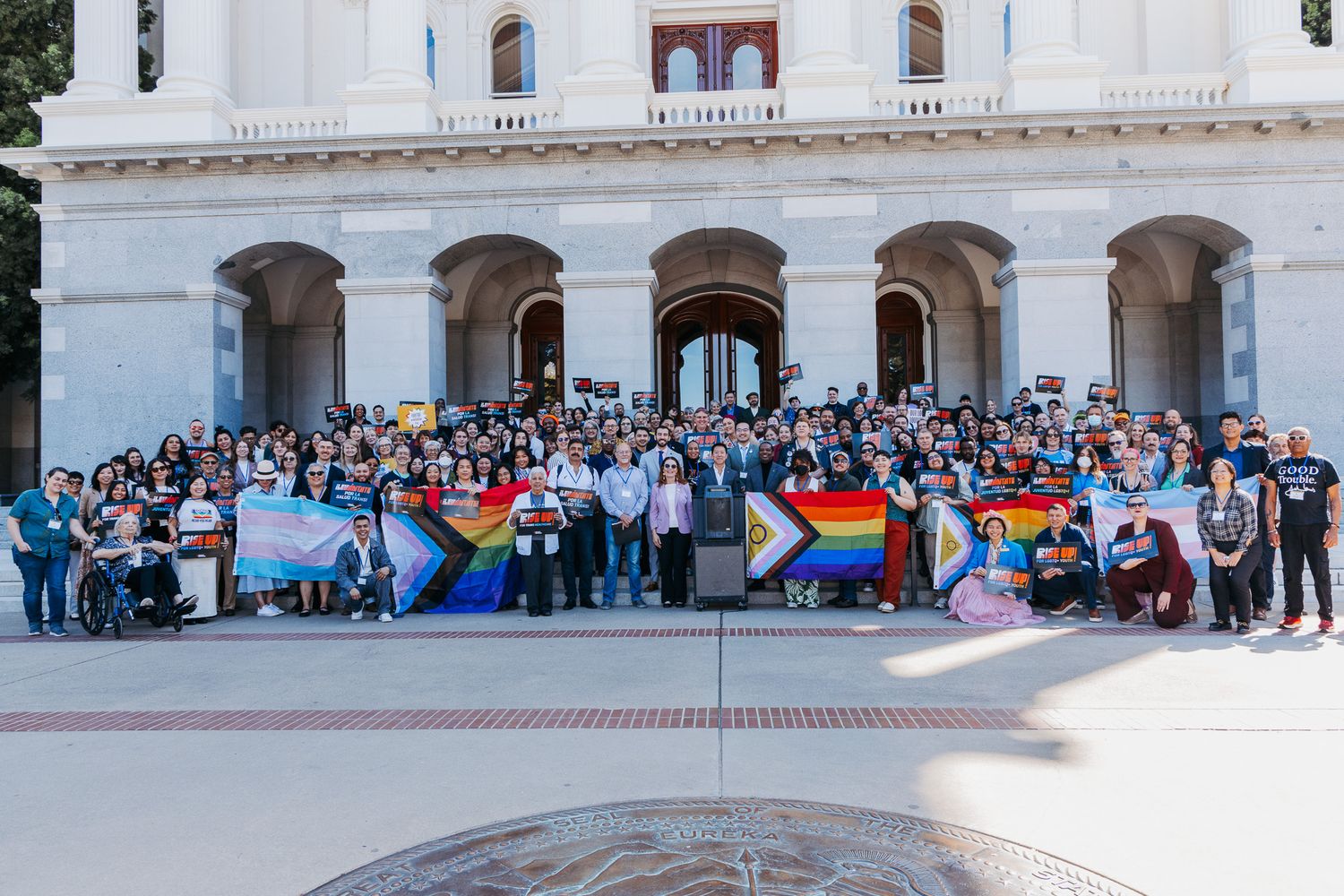 A crowd of attendees sits on steps in front of a white columned building during an outdoor gathering.