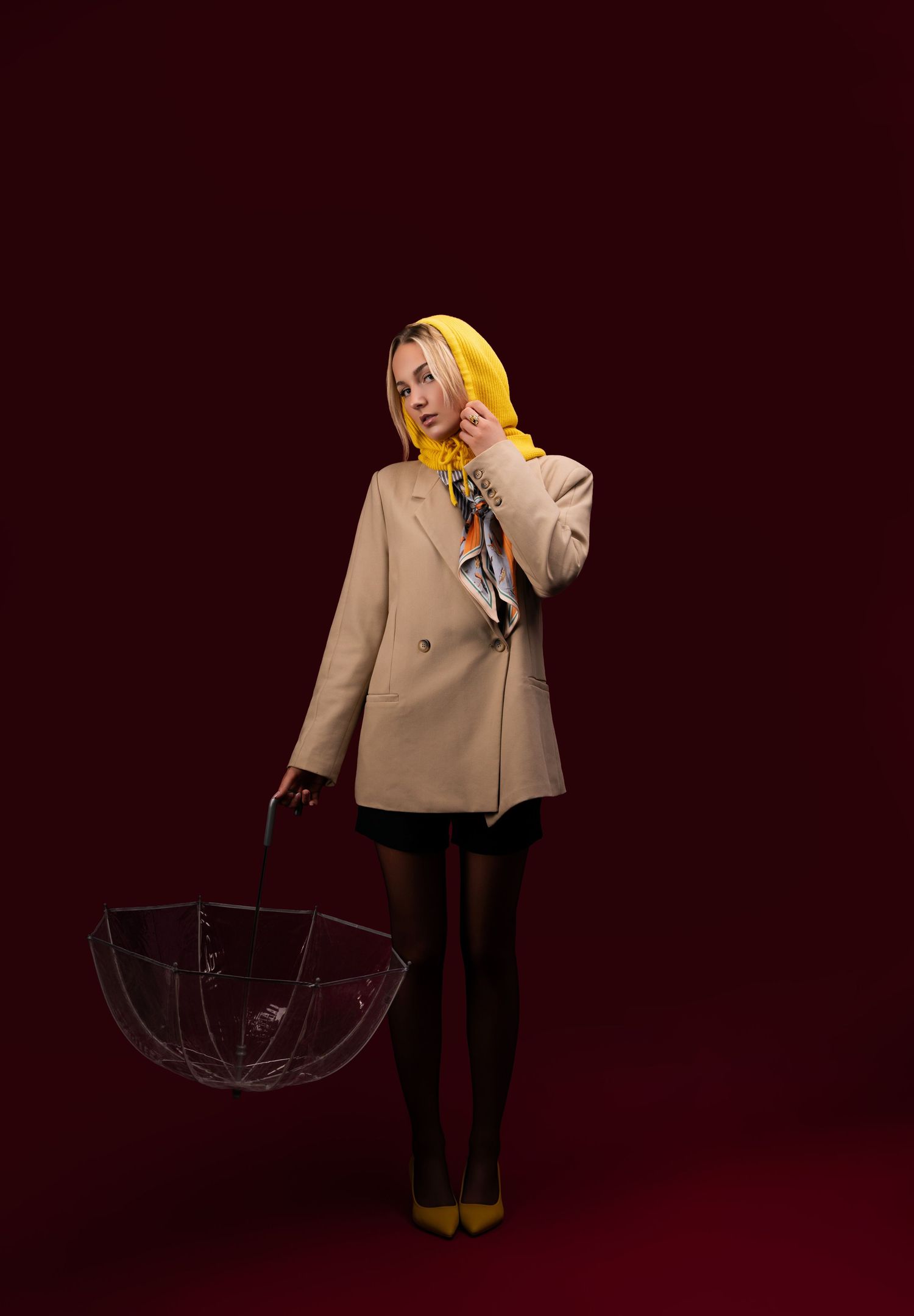 Stylish figure in beige coat and yellow boots poses with black umbrella against deep burgundy backdrop.