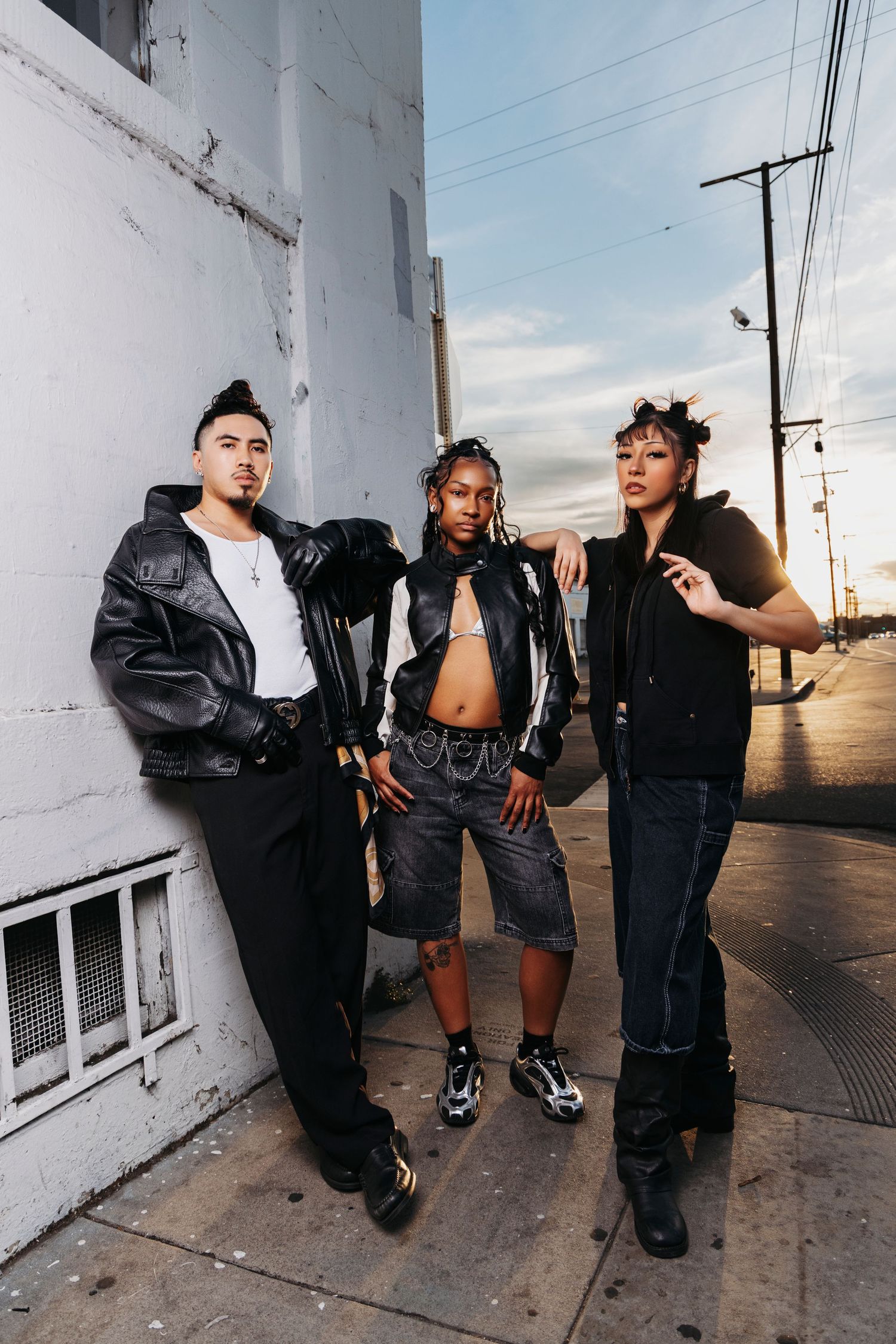 A group poses against a white wall in an urban setting wearing black leather jackets and casual streetwear.
