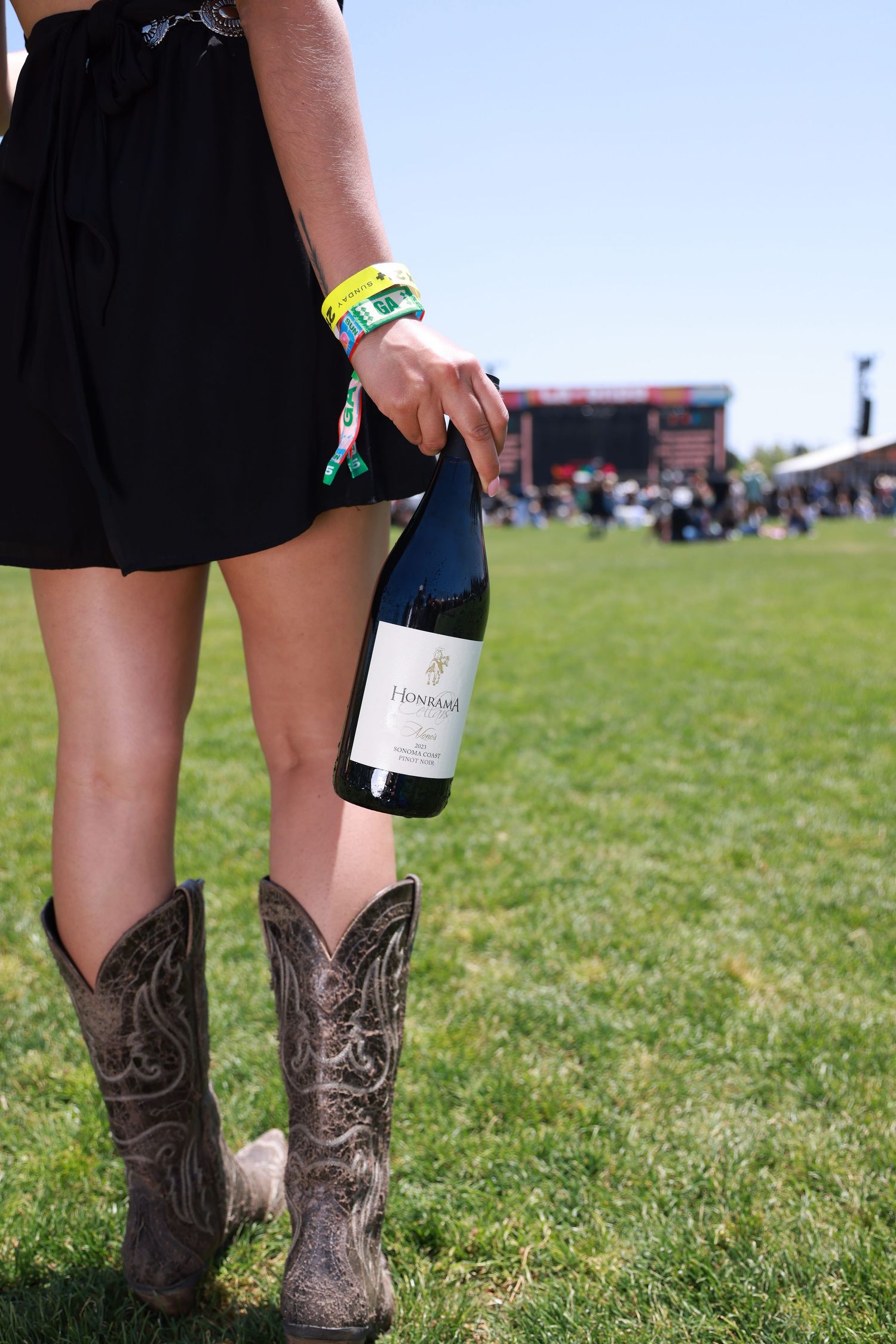 Cowboy boots stand out against green grass at an outdoor festival with a wine bottle visible.