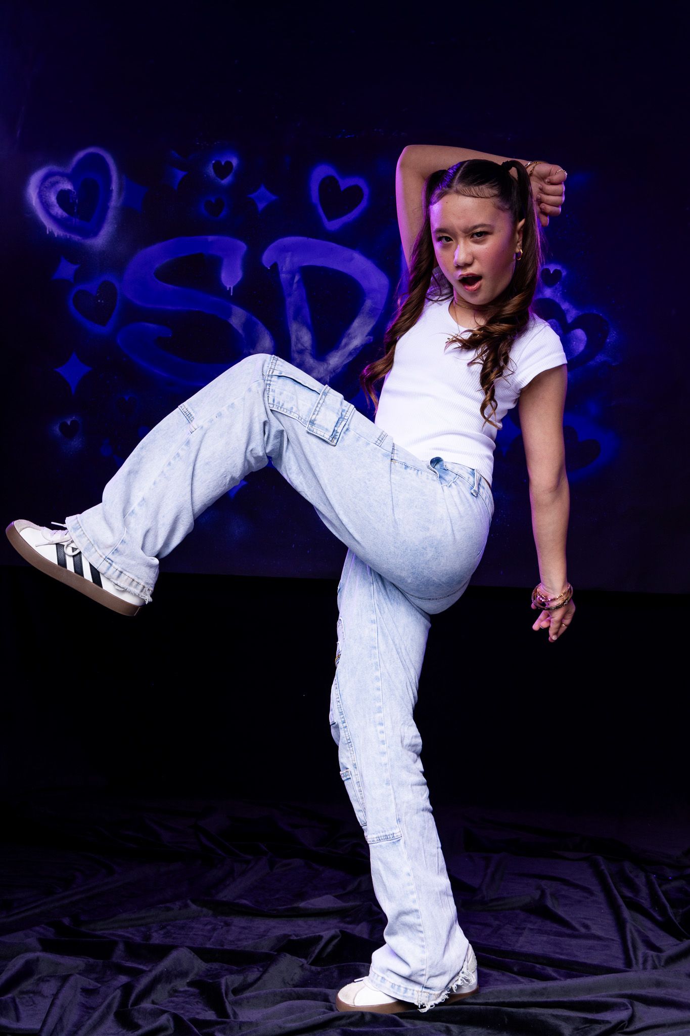 Young dancer in white crop top and light blue jeans strikes a dynamic hip-hop pose against a dark blue graffiti backdrop.