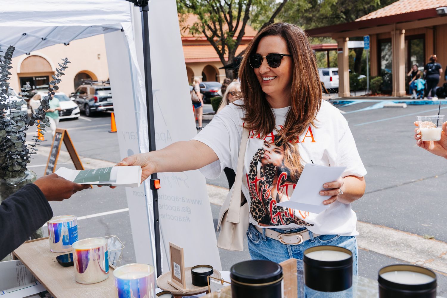 Woman in white tee smiles while handing item at outdoor market candle vendor booth on sunny day.