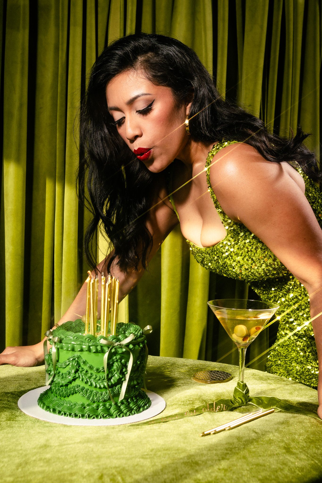 Person in sparkling green dress elegantly blowing out birthday candles on a dark green frosted cake.
