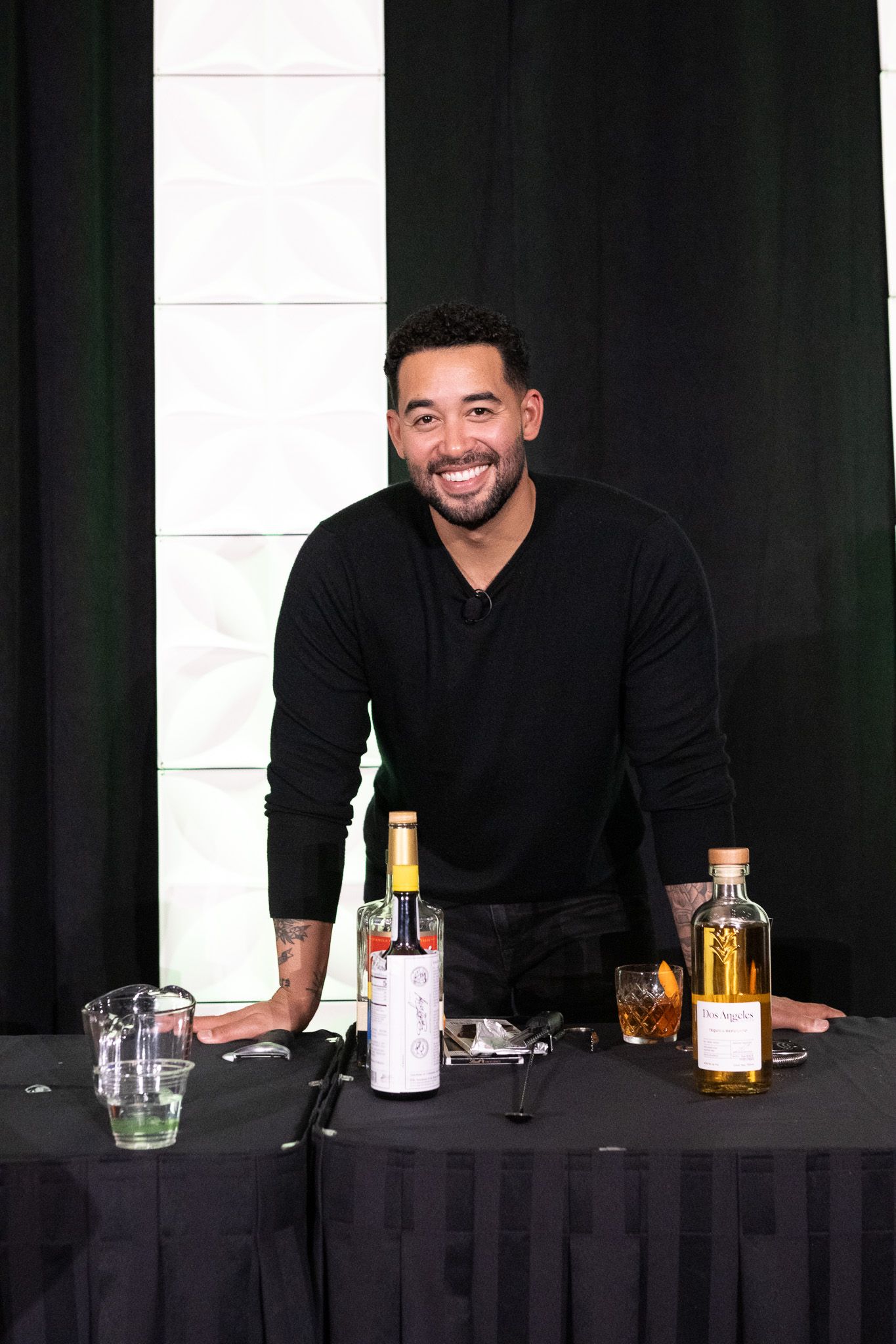 A black-clad person smiles while standing behind a table with various bottles and glasses at what appears to be an event.