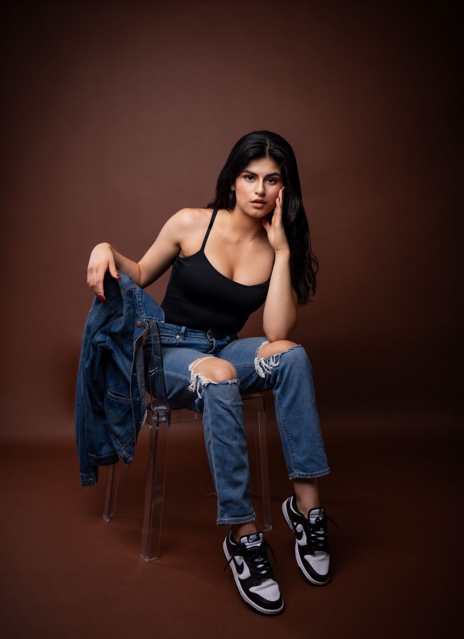Model in denim and black tank top poses on wooden stool against dark brown background in fashion photoshoot series.