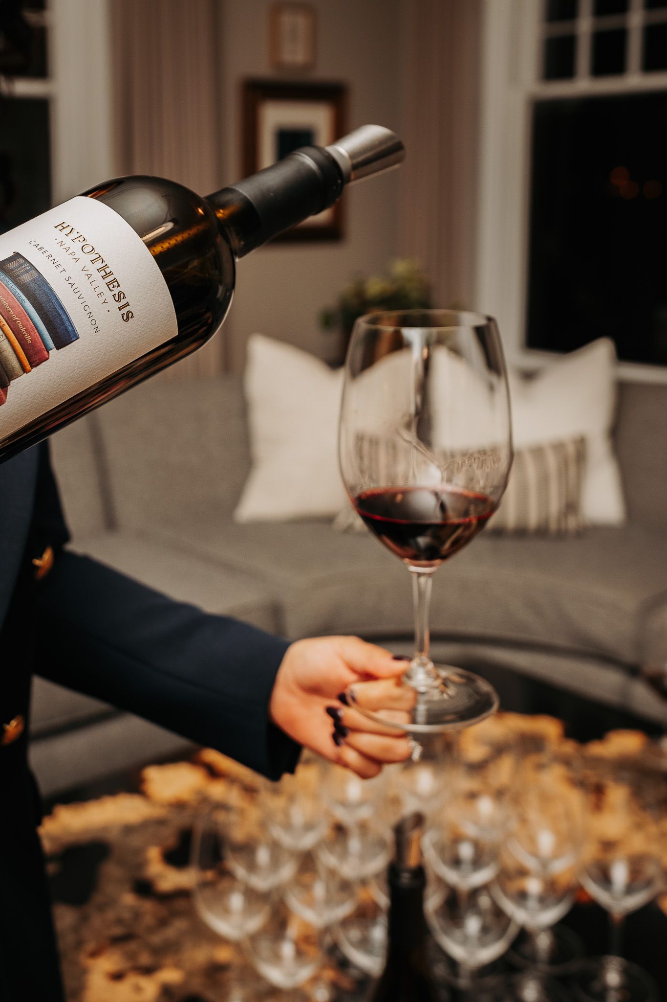 Red wine being poured into elegant wine glasses at an evening gathering with soft lighting and appetizers.