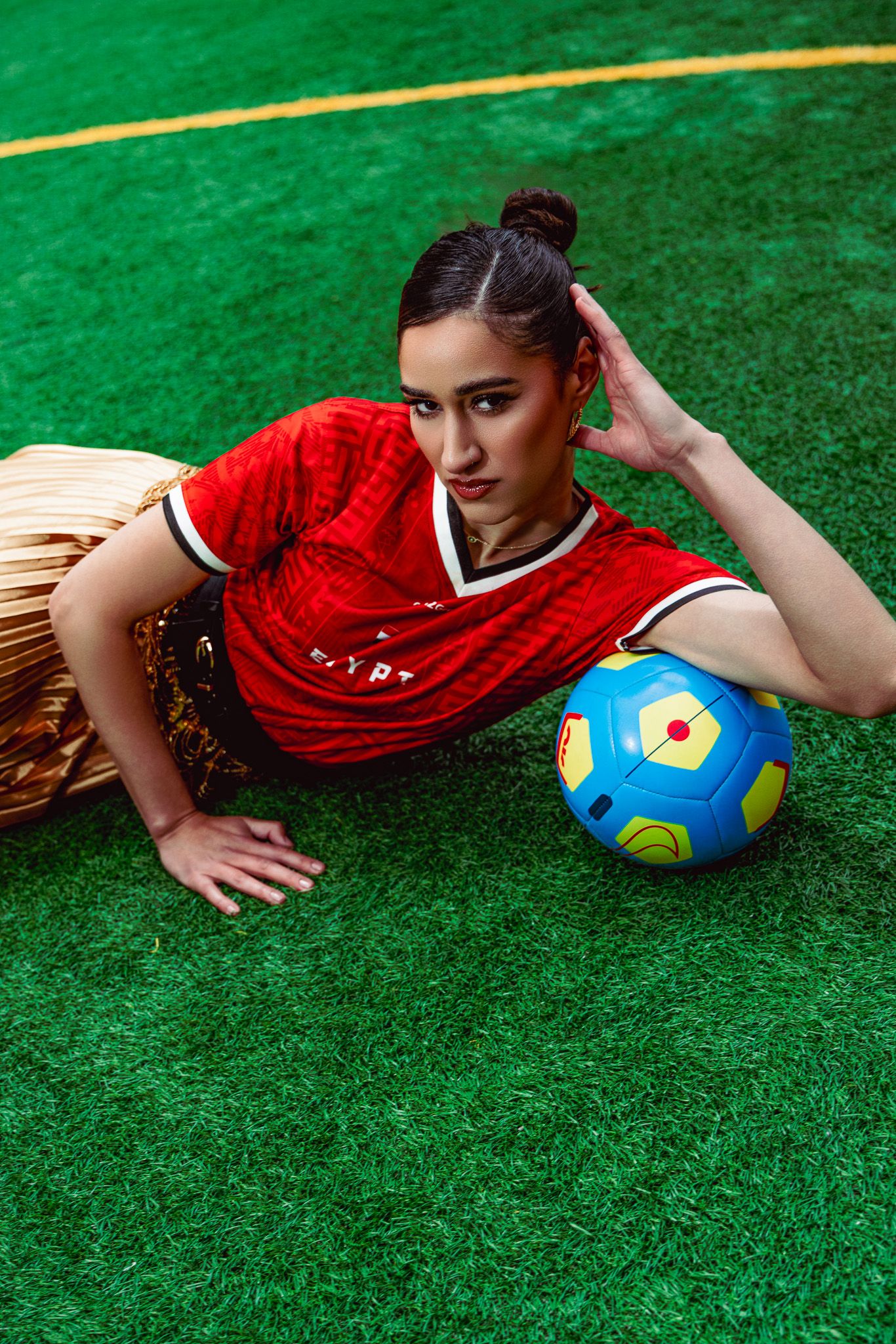 Woman in red soccer jersey poses on green turf field with colorful blue soccer ball.
