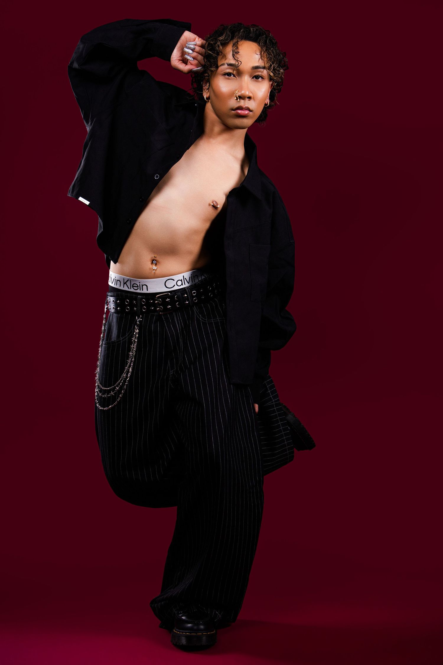 Young man posing shirtless in black jacket, pinstripe pants with chain belt and Calvin Klein underwear on dark red background.