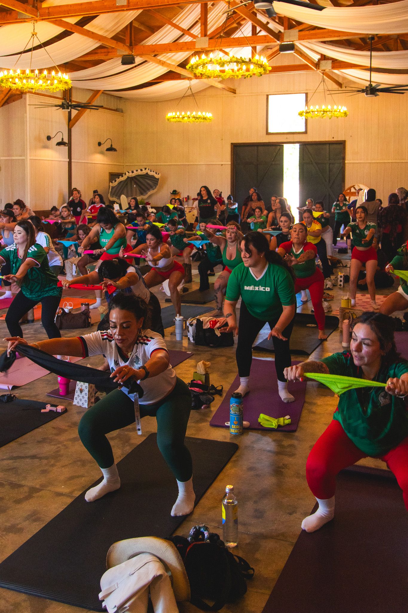 Large group fitness class with women doing yoga poses on mats in a decorated hall with chandeliers.