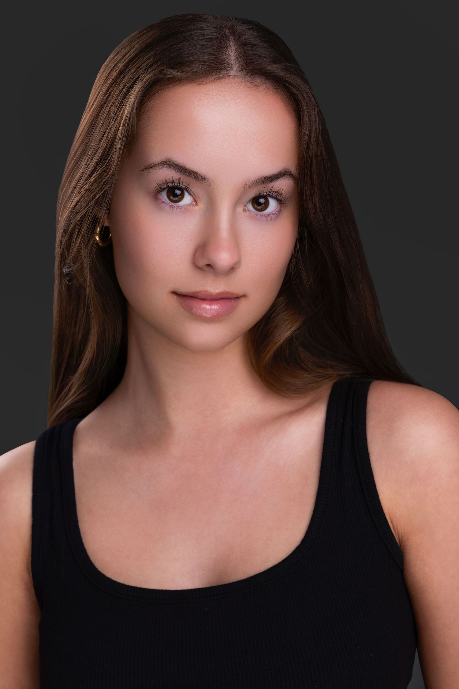 Professional portrait photo with dark background showing a person in a black tank top with long brown hair and neutral makeup.