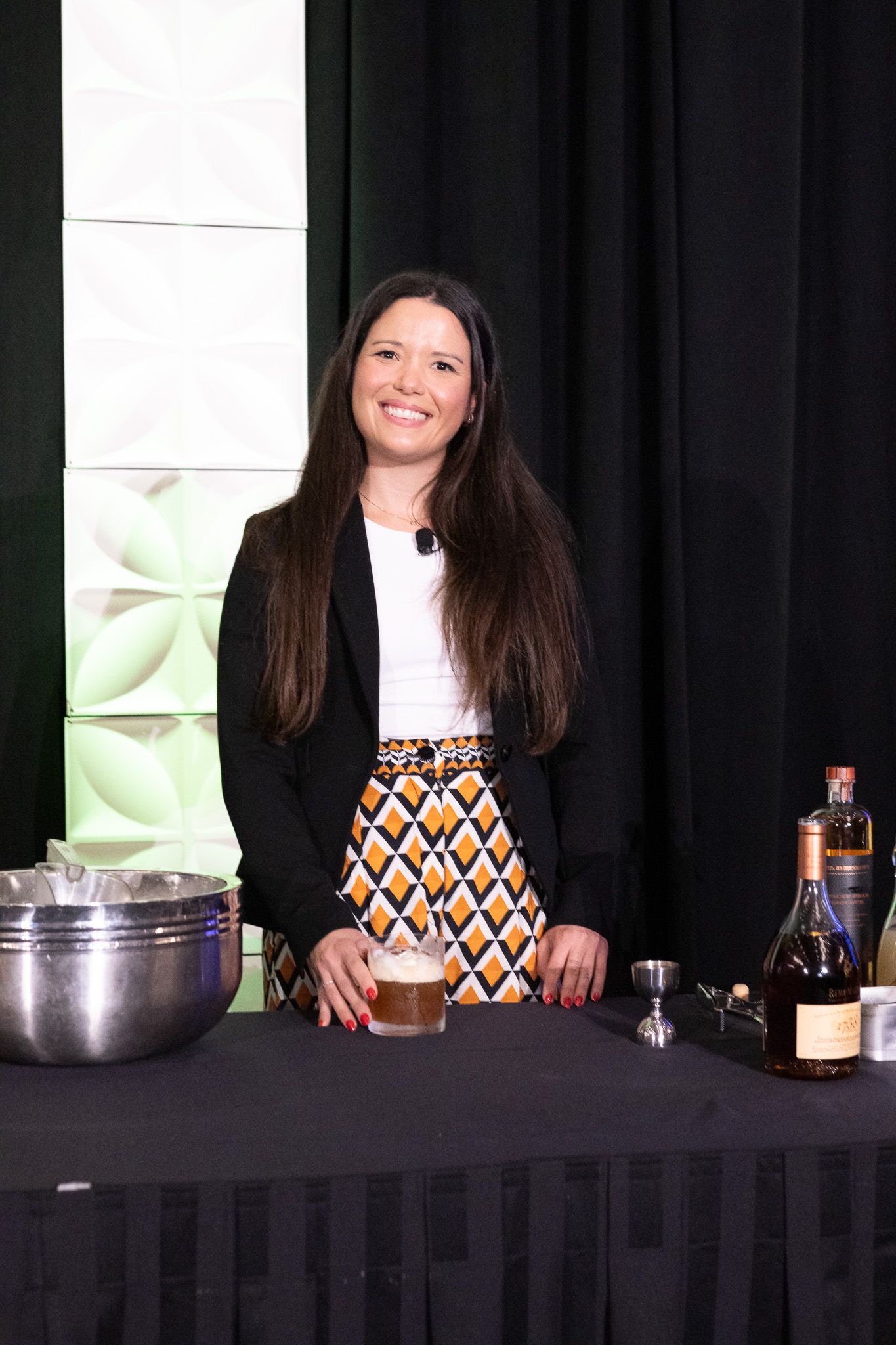 Professional bartender in black blazer and patterned skirt stands behind table with cocktail mixing equipment and bottles.