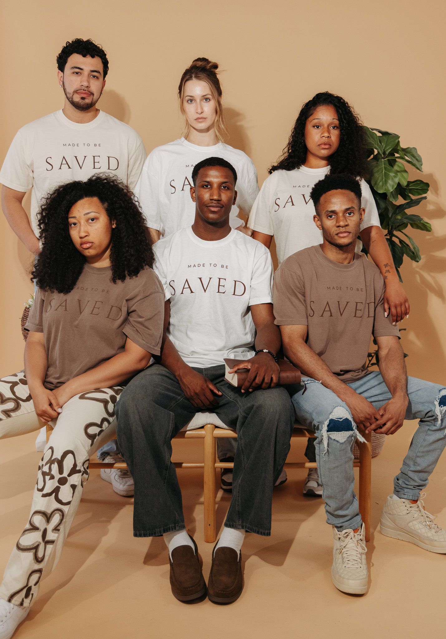 Group photo of people wearing beige and white t-shirts with 'SAVED' text sitting together on wooden bench.