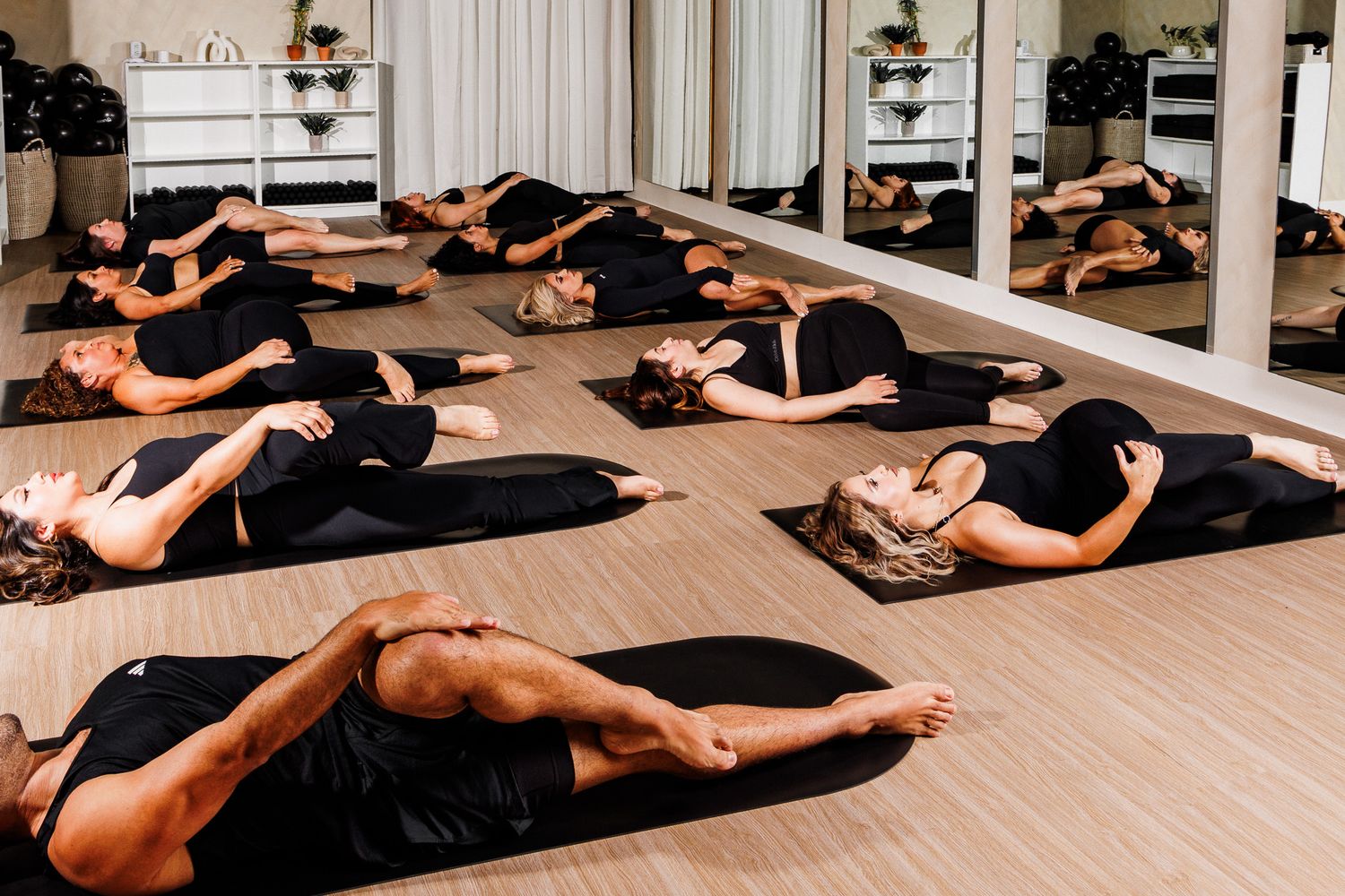 Group Pilates class lying on black mats in a bright mirrored fitness studio.