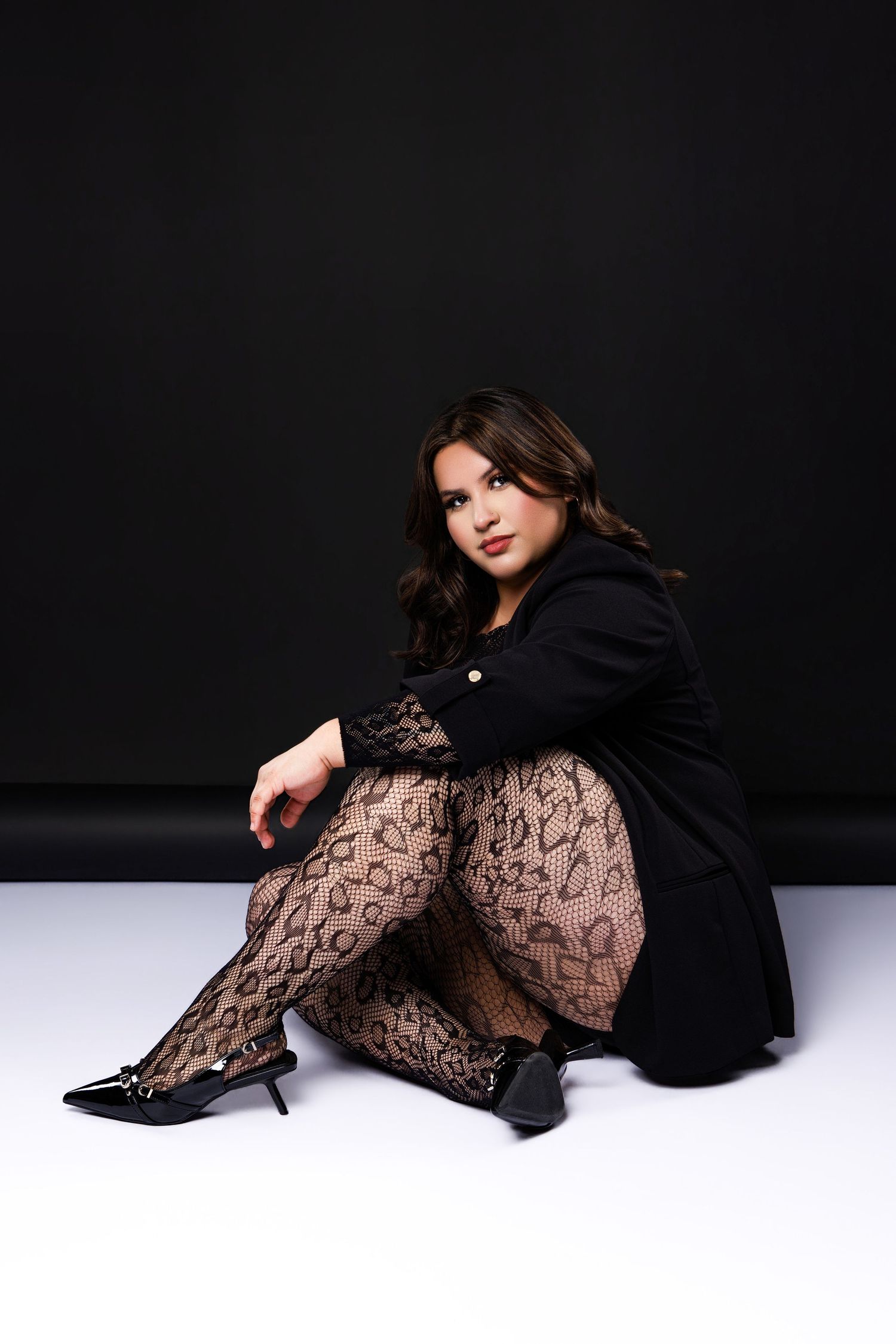 Woman in black blazer and lace tights sitting on floor against dark background wearing heeled pumps.