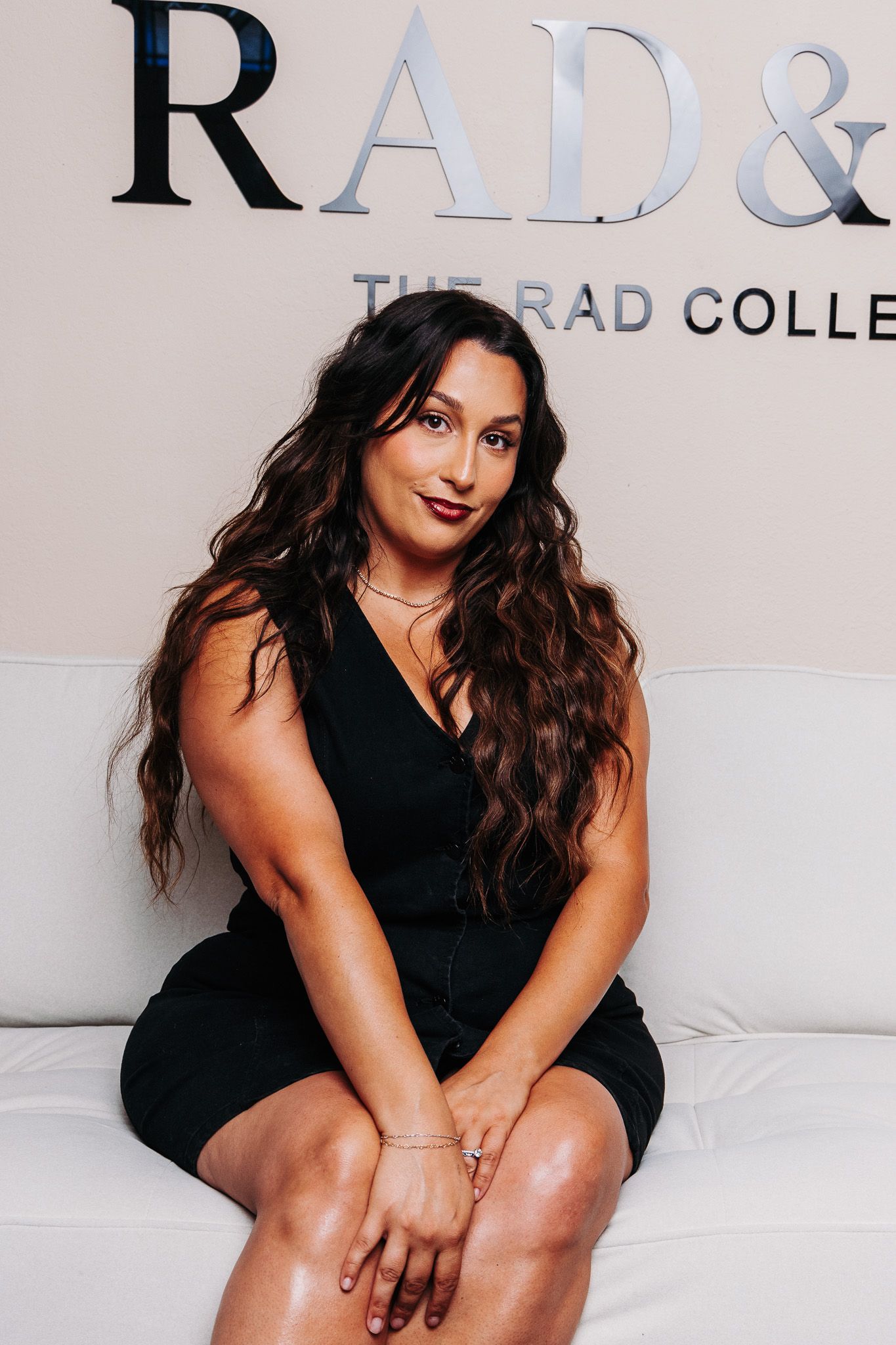 Woman in black dress sits confidently in front of RAD & Co. collection sign, with long wavy hair and elegant jewelry.