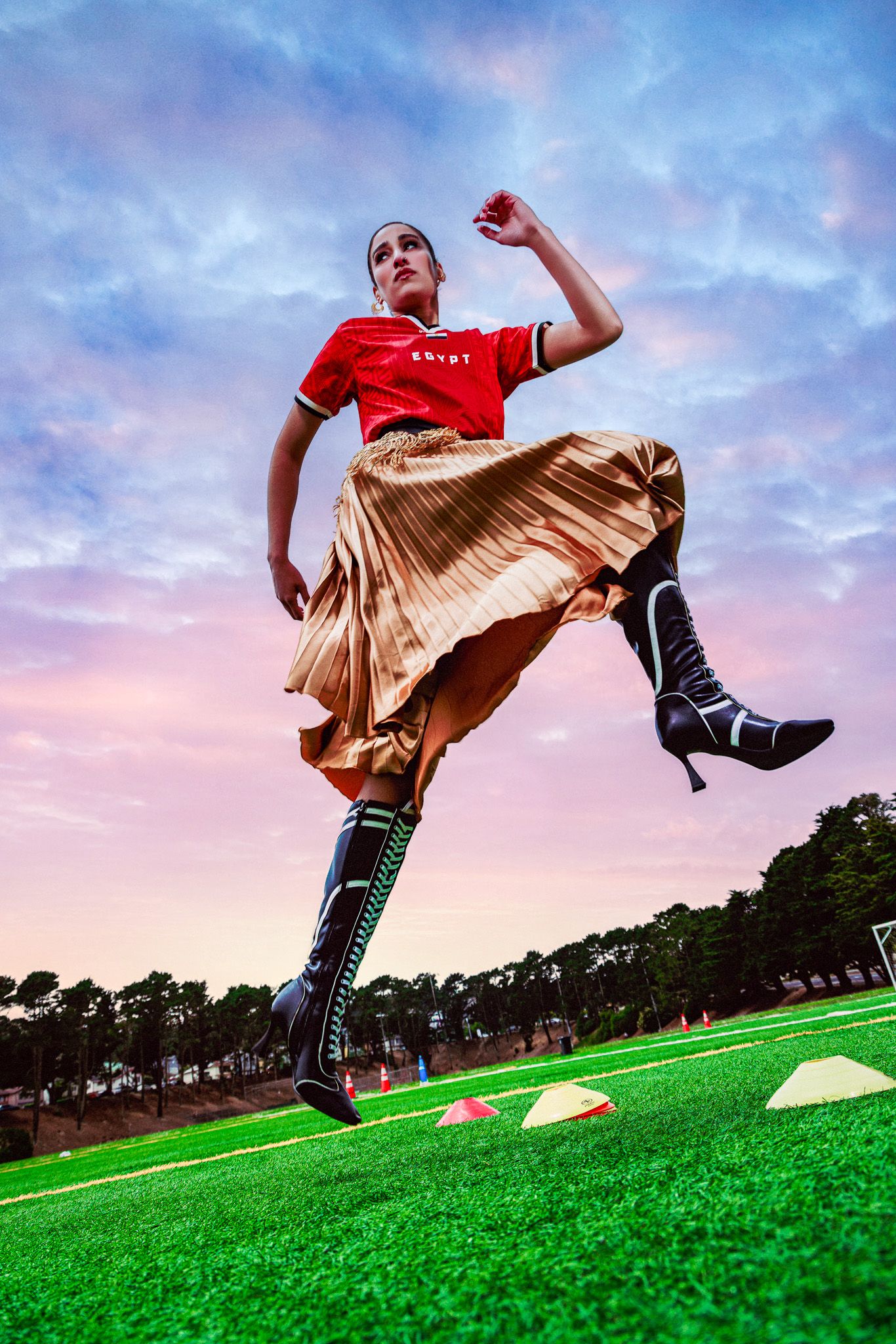 Woman in red top, gold pleated skirt, and black boots jumping on a green soccer field at sunset.