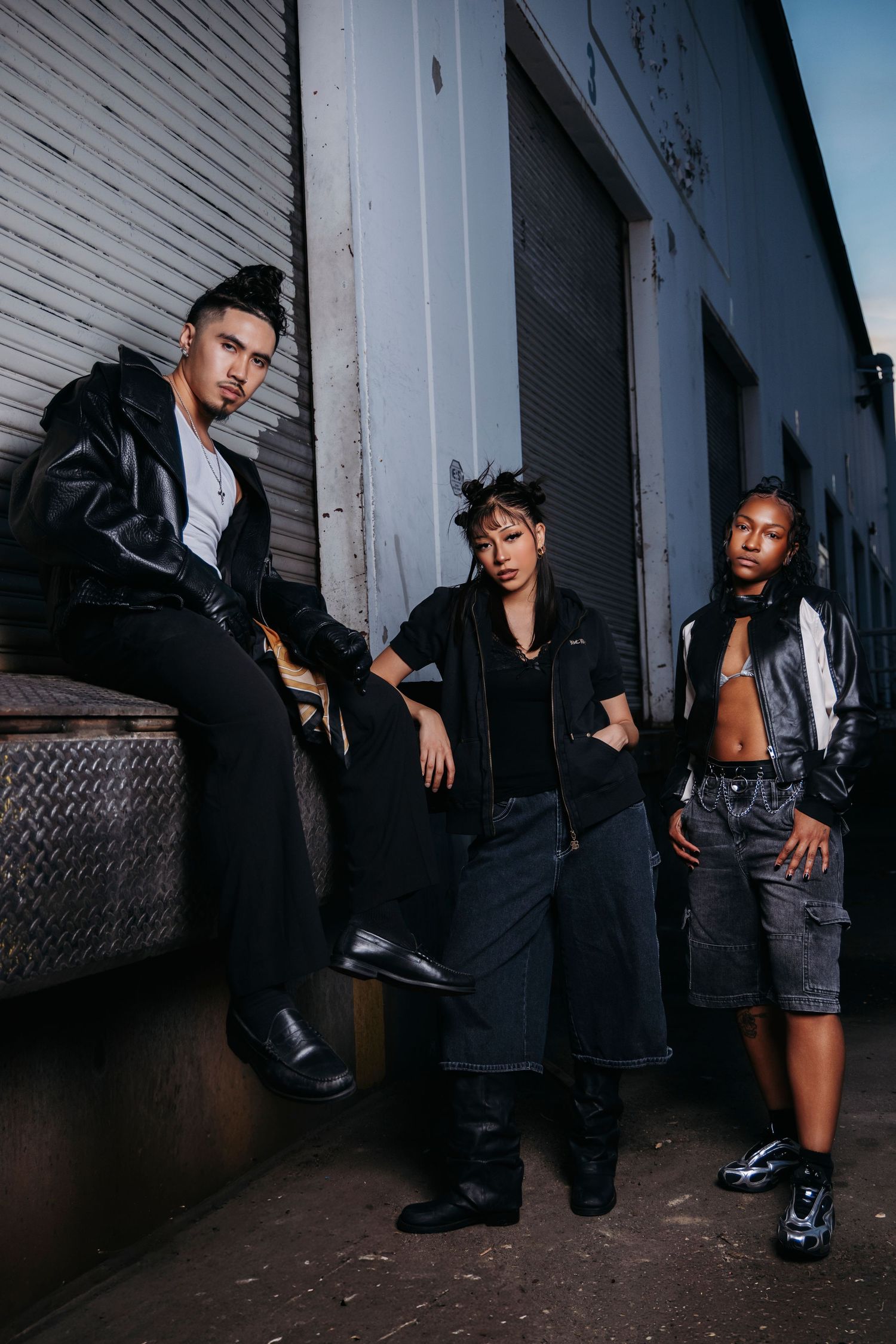 Fashion models in dark outfits pose dramatically against industrial backdrop in moody lighting.