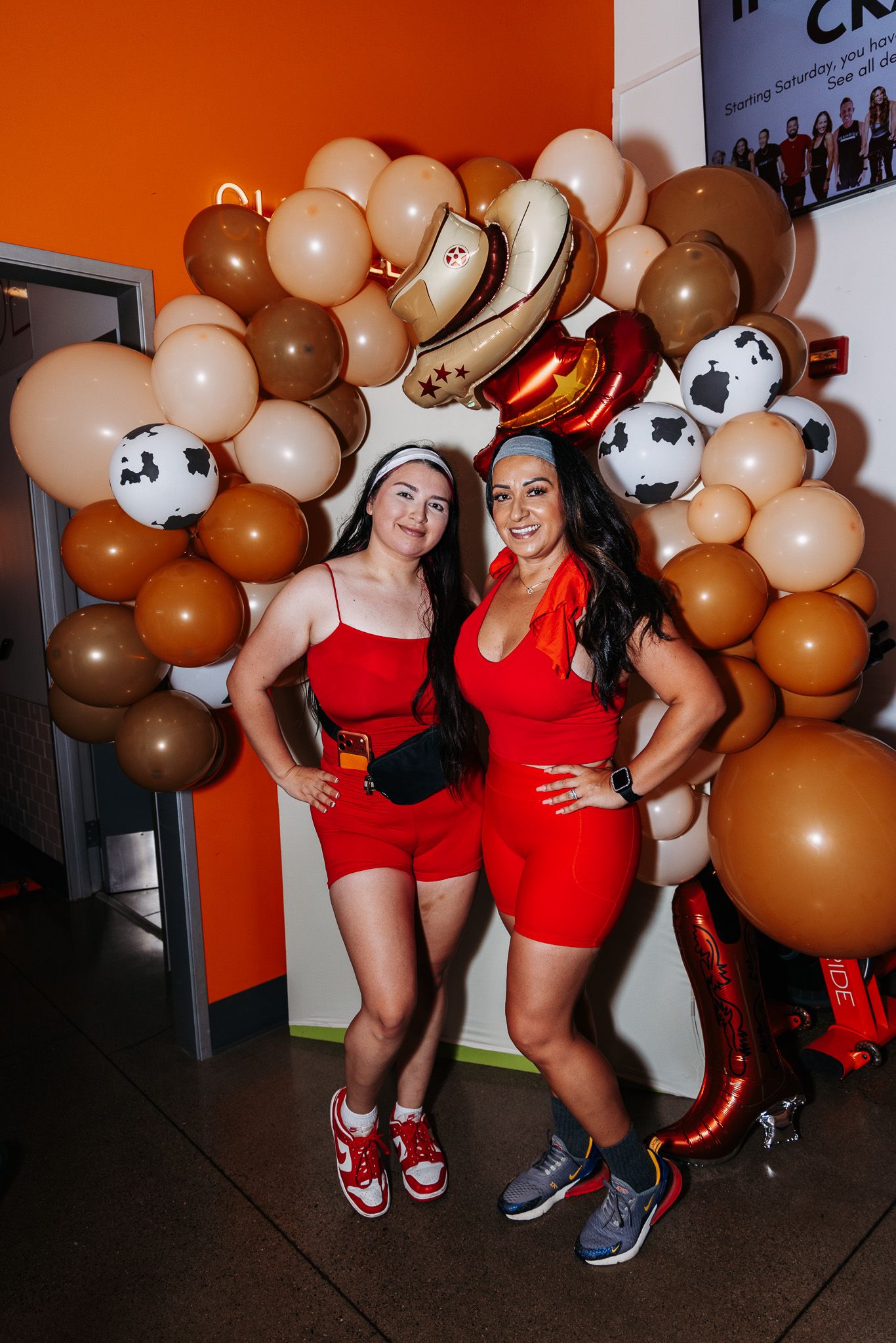 Two people in matching red outfits pose in front of a brown and beige balloon arch at a party.