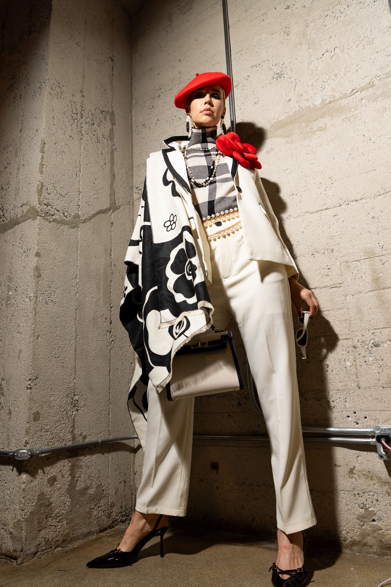 Stylish outfit featuring white pants, black and white patterned jacket, red beret and neck scarf against concrete wall.
