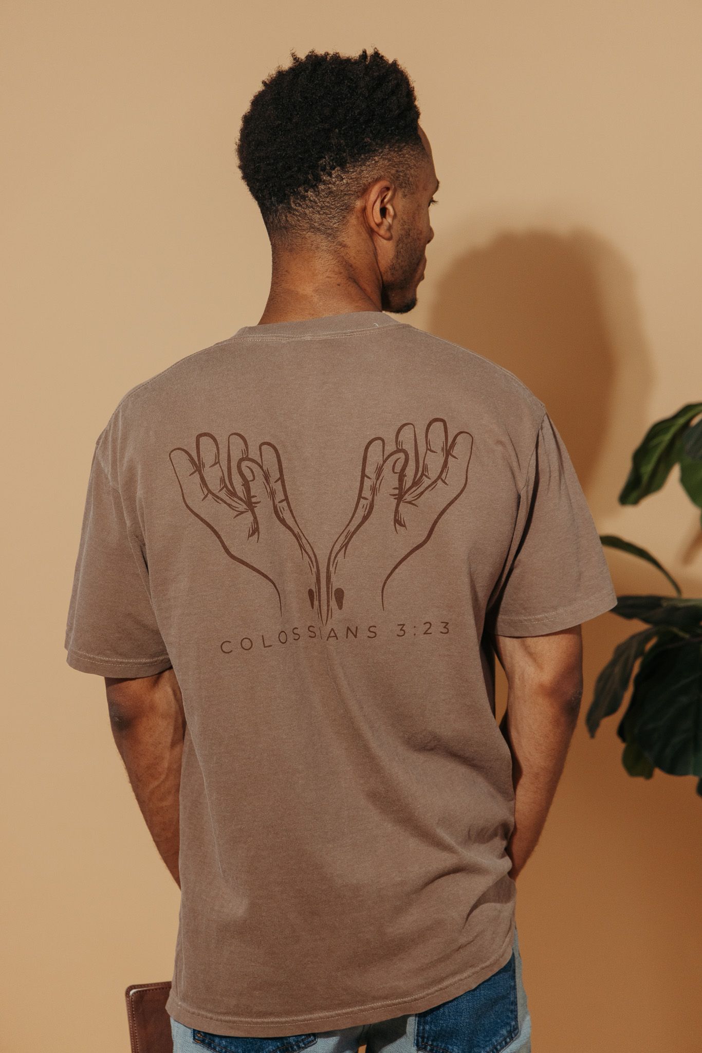 Back view of person wearing brown t-shirt with hand illustrations and scripture reference Colossians 3:23.