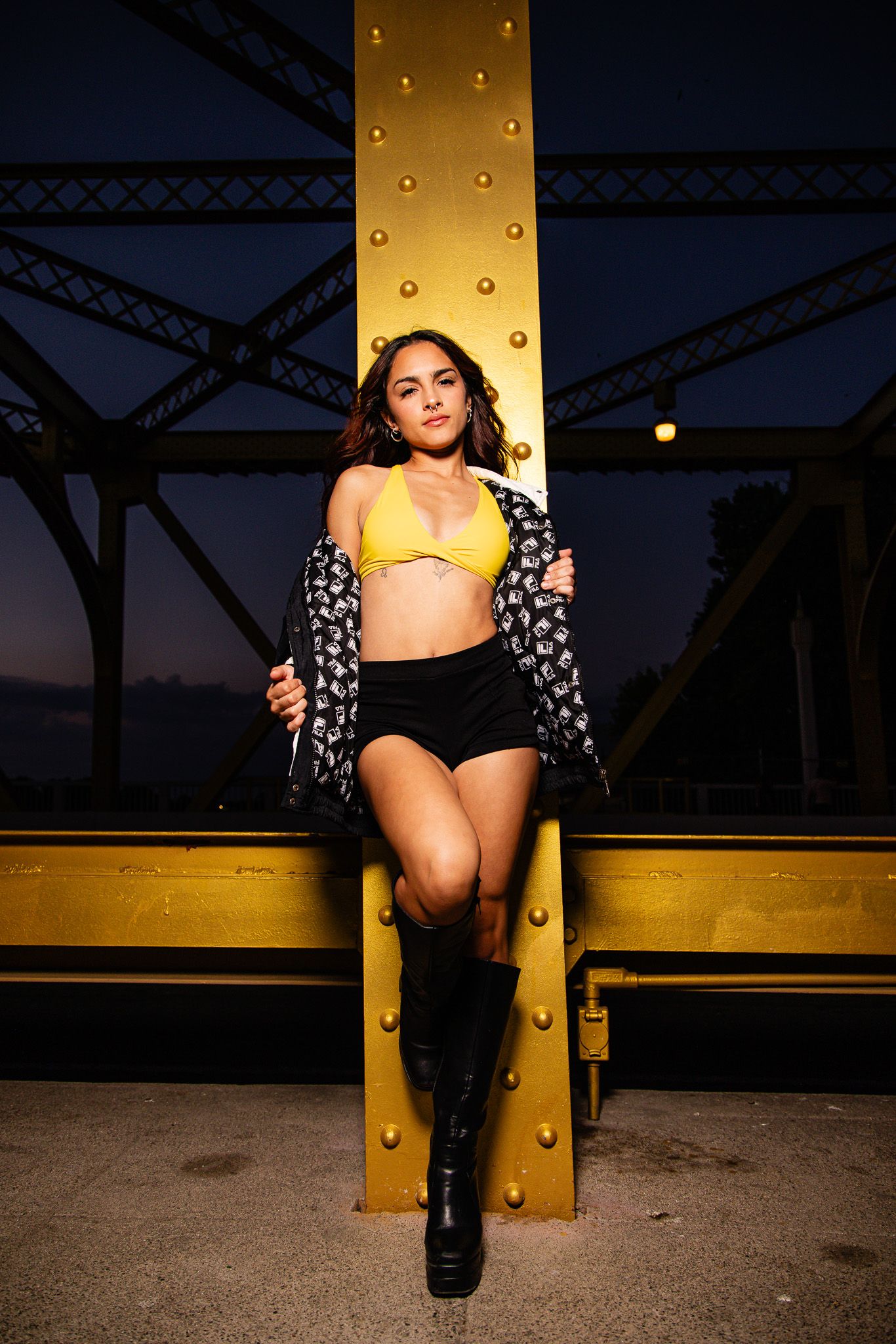 A person in a dark floral print jacket poses at dusk against an industrial bridge structure with dramatic lighting.