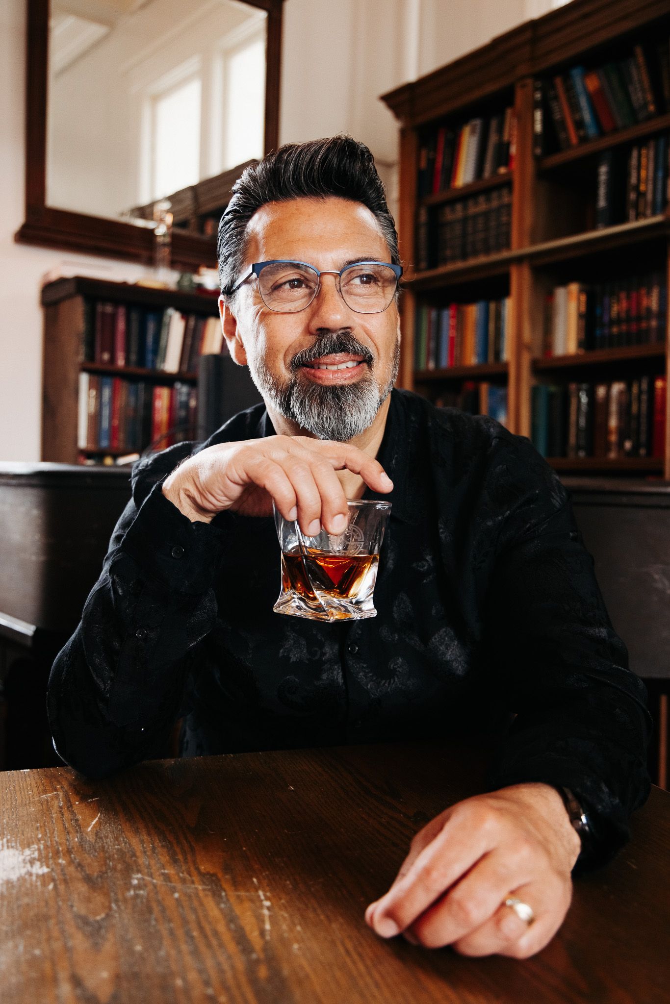 Someone enjoying a glass of whiskey or bourbon while seated at a wooden bar with bookshelves visible in the background.