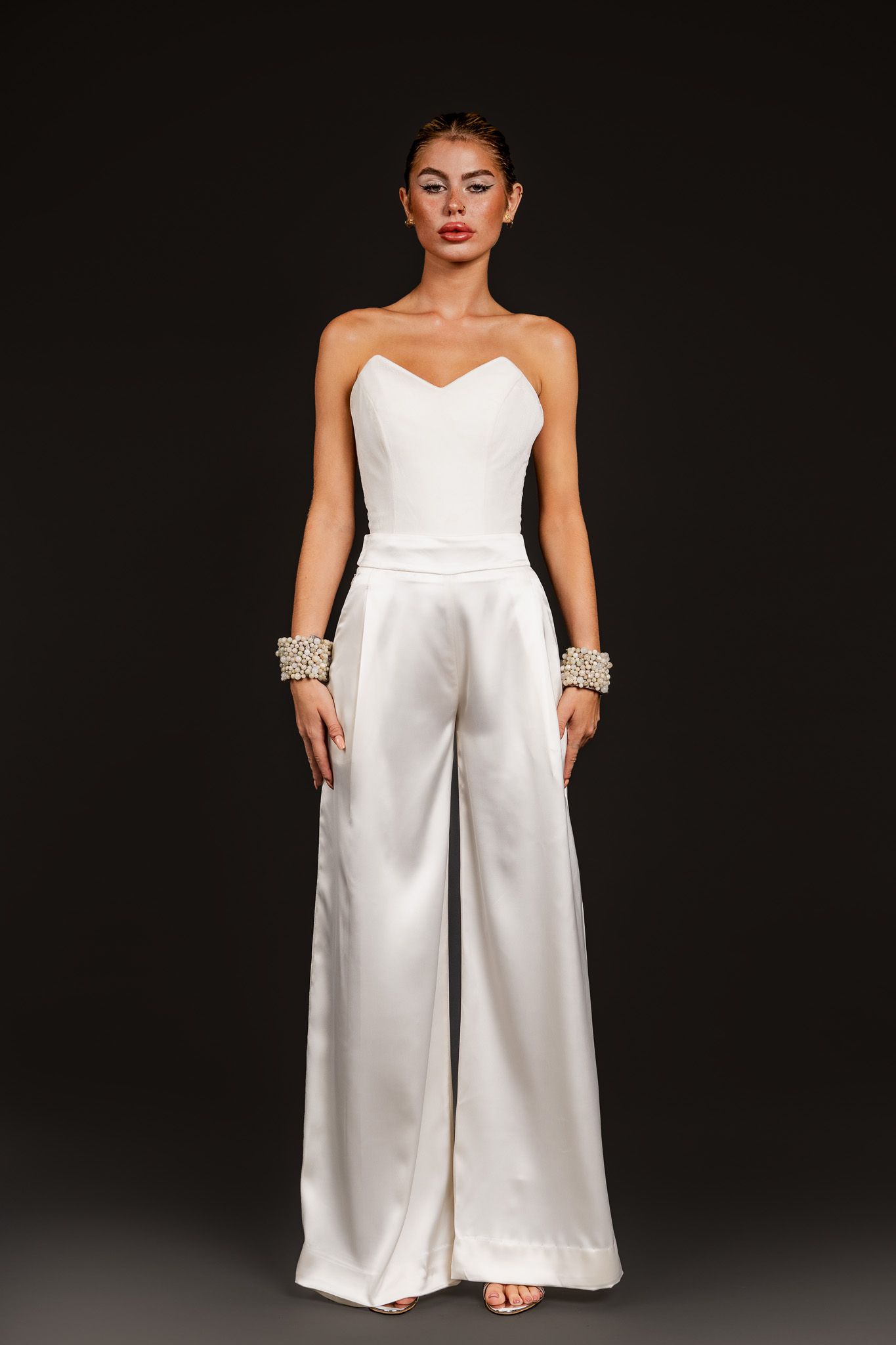 Woman wearing a white strapless satin wide-leg jumpsuit with crystal cuff bracelets on a dark background.