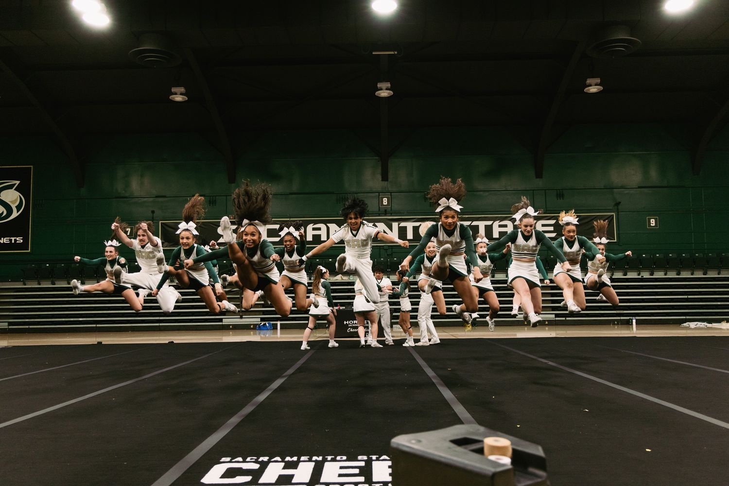 Sacramento State cheerleaders performing a dynamic jump routine on a green and black gym floor during practice.