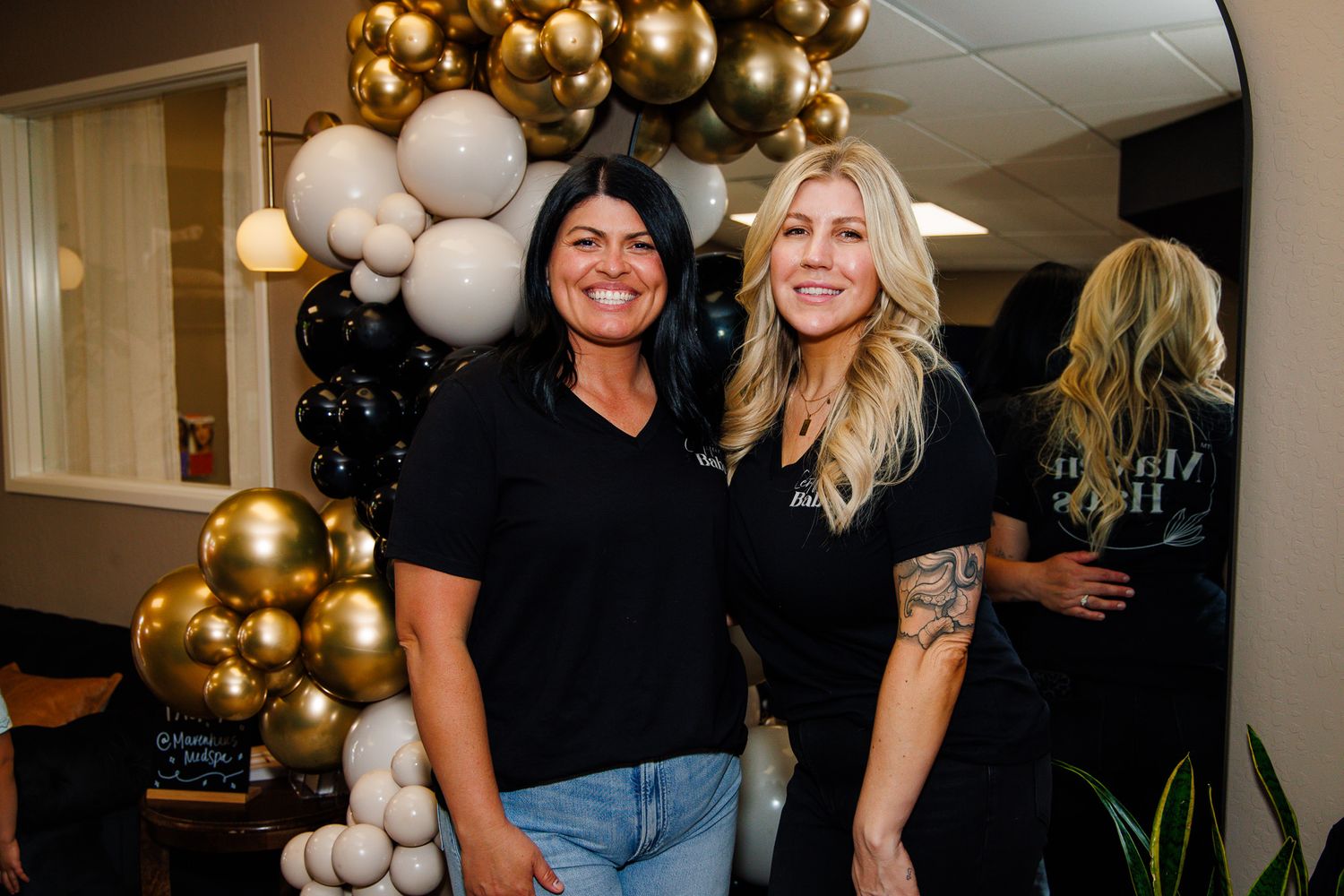 Two women in black shirts smile together in front of gold and black balloon arch display.