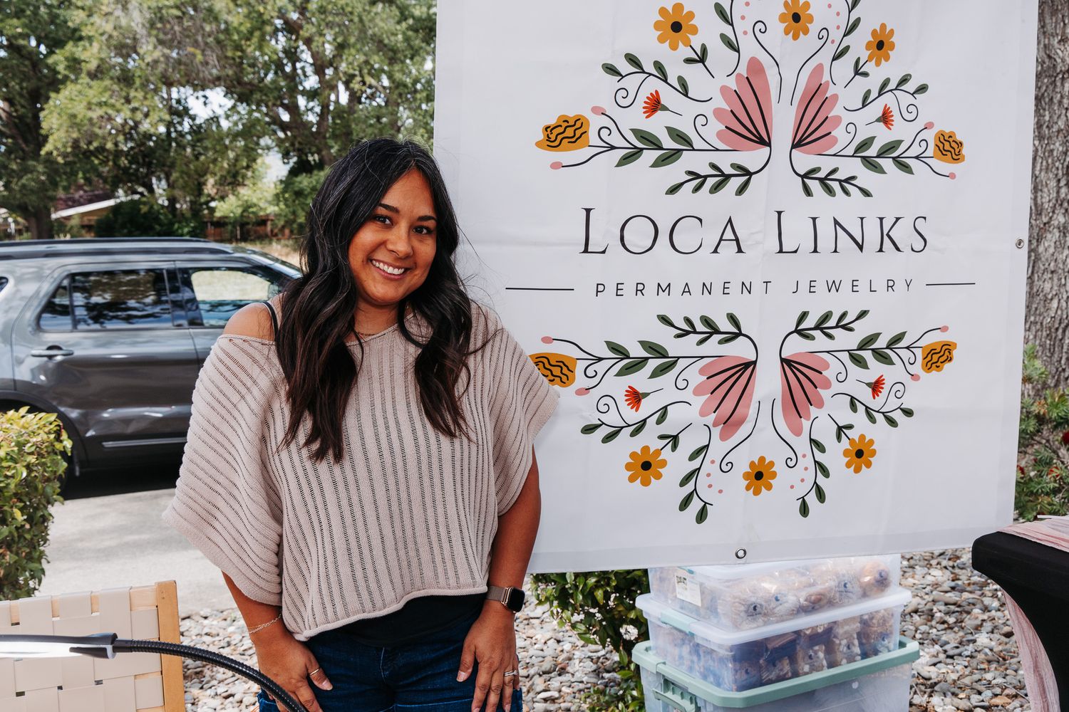 Woman smiling beside Loca Links Permanent Jewelry sign at outdoor market booth.