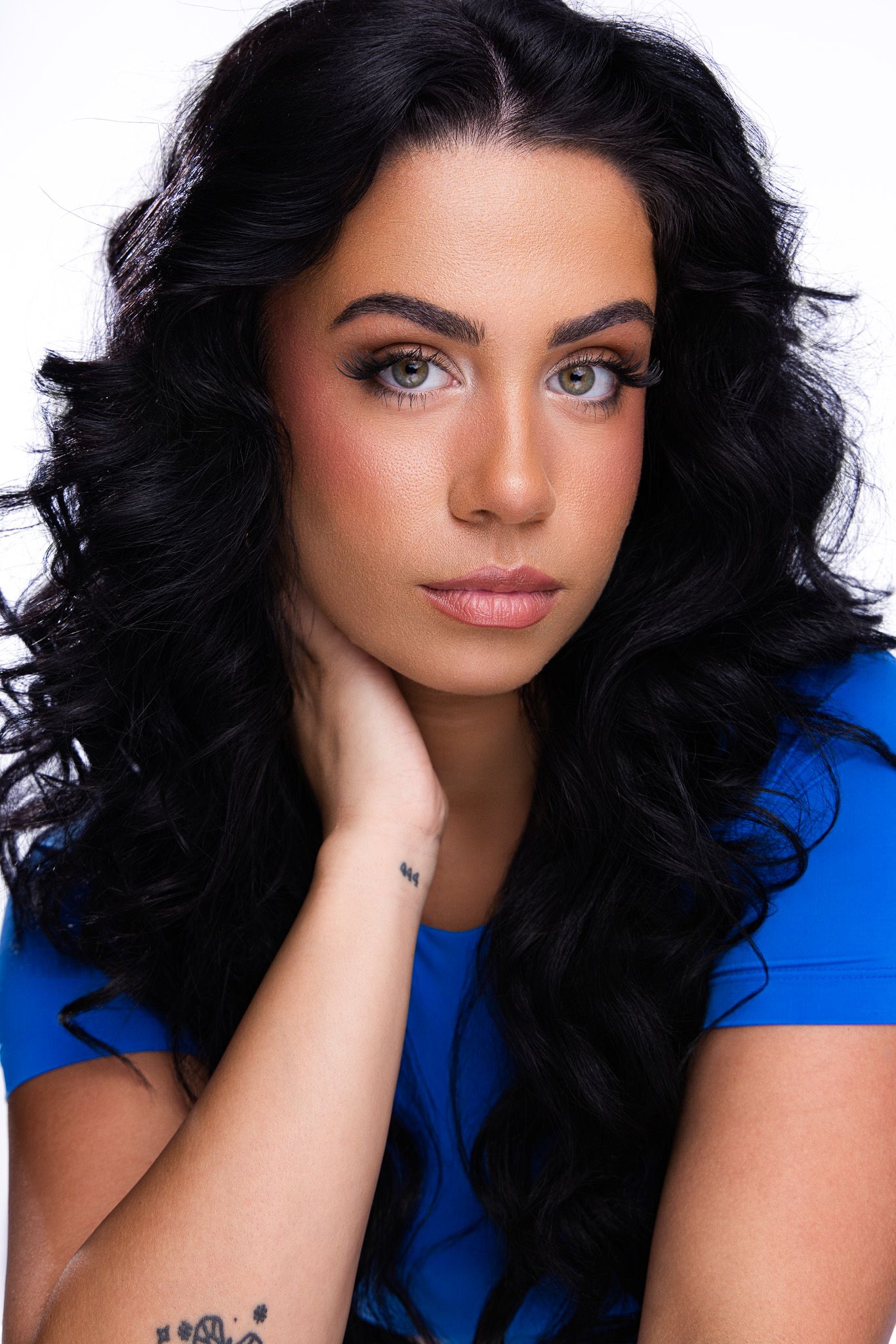 Young woman with long curly black hair, green eyes, blue top, and wrist tattoo poses against white background.