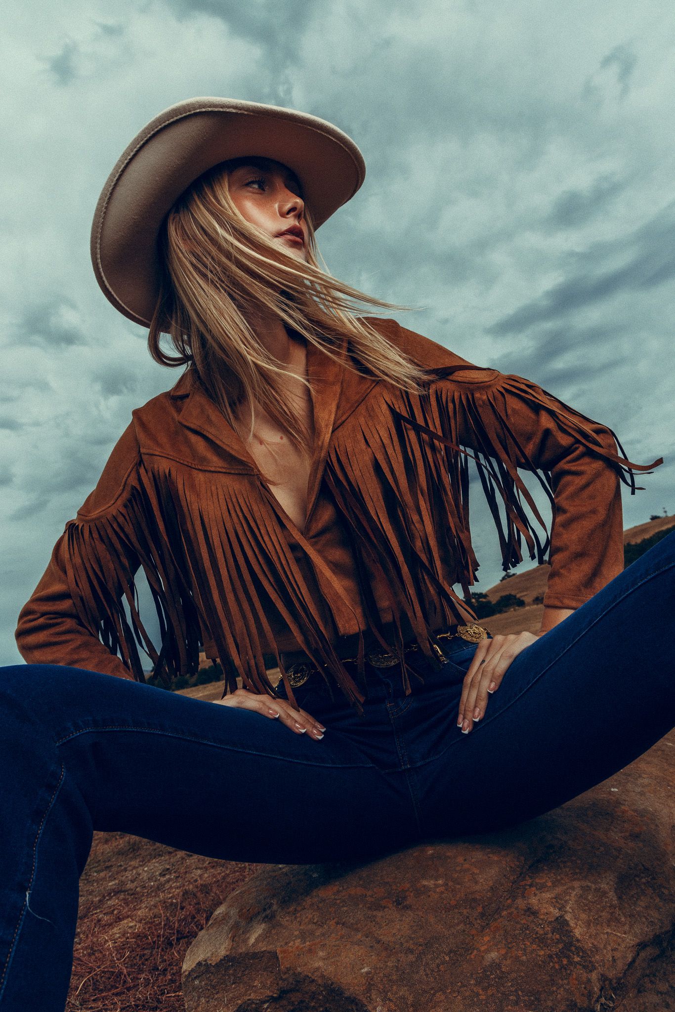 A dramatic fashion shot showing a brown fringe leather jacket against a moody cloudy sky in the desert.